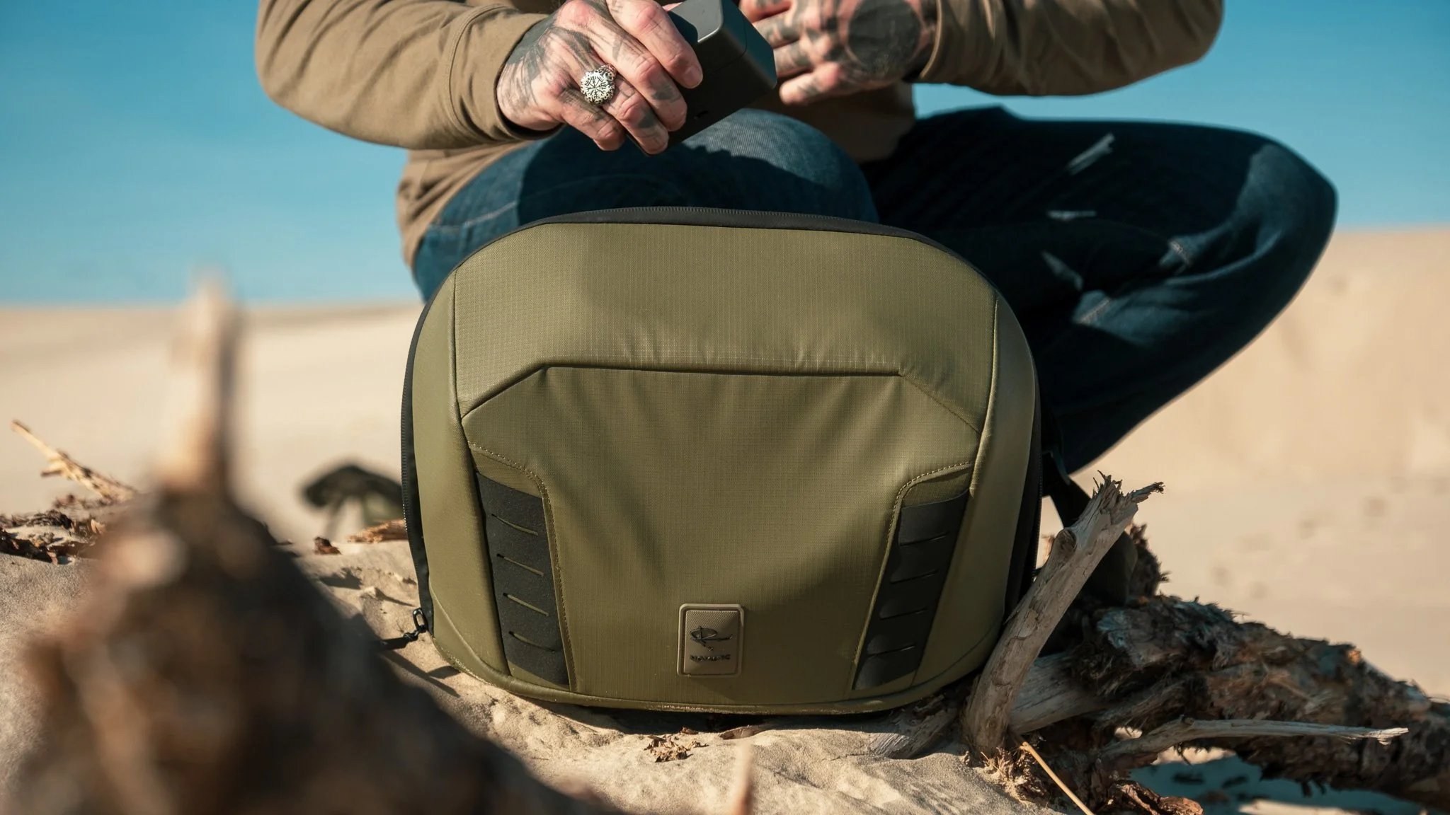 Person sitting on sand beach with a green backpack nearby, holding a camera or phone, with the ocean in the background.