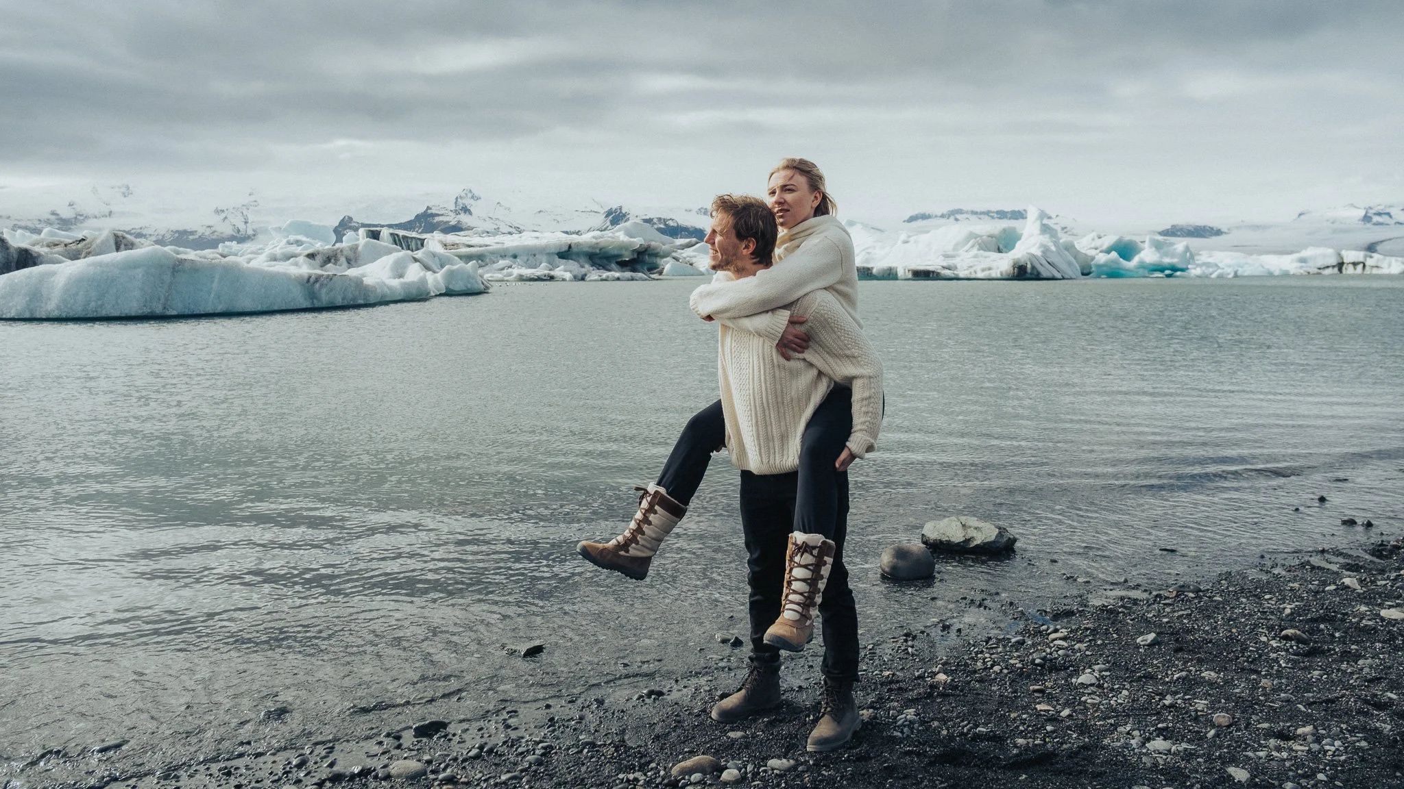 A couple standing by icy waters with glaciers and mountains in the background, the woman is piggybacking on the man, both wearing warm sweaters and boots, in a cold, Arctic landscape.