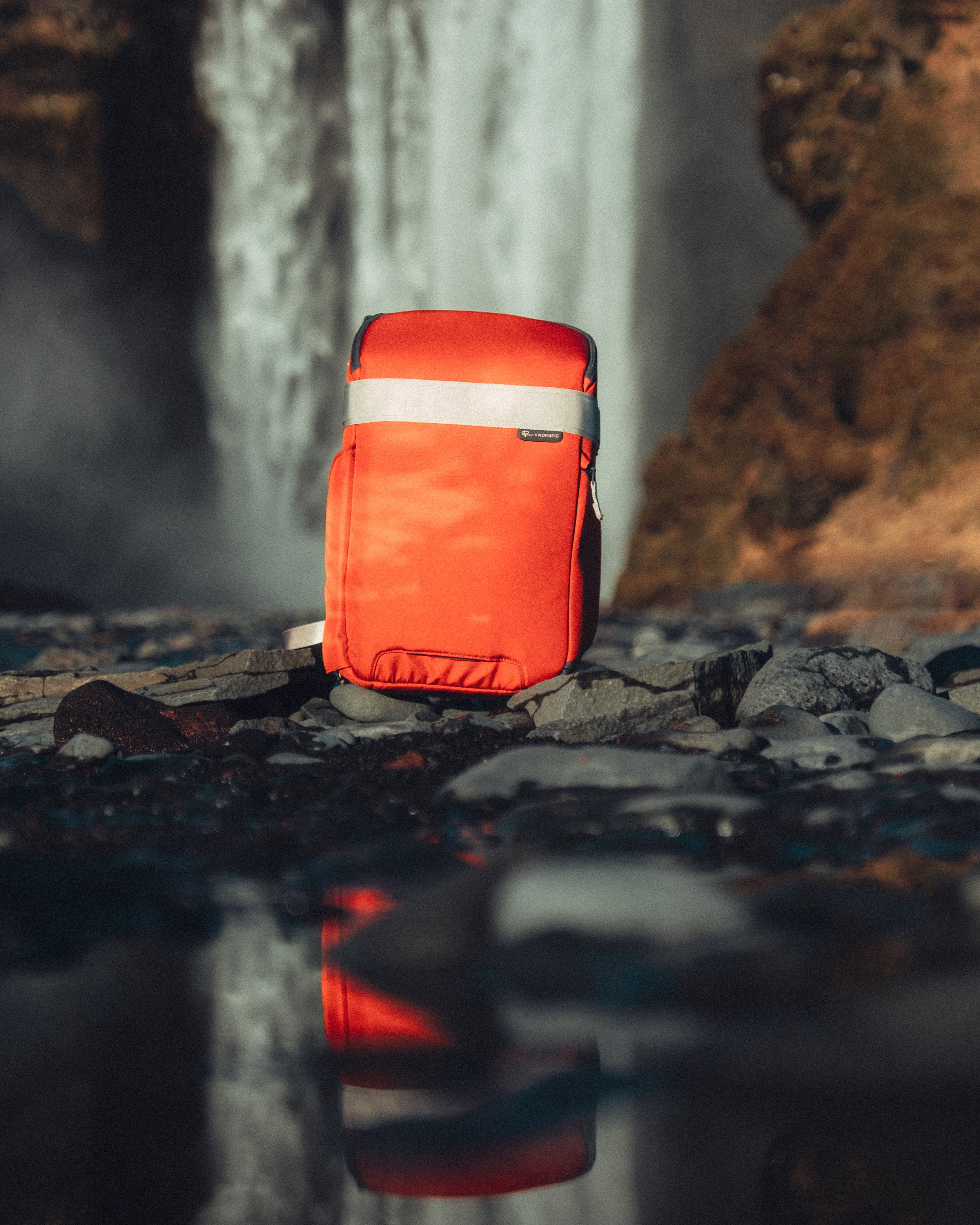 An orange travel backpack on rocky ground near water with a waterfall in the background.