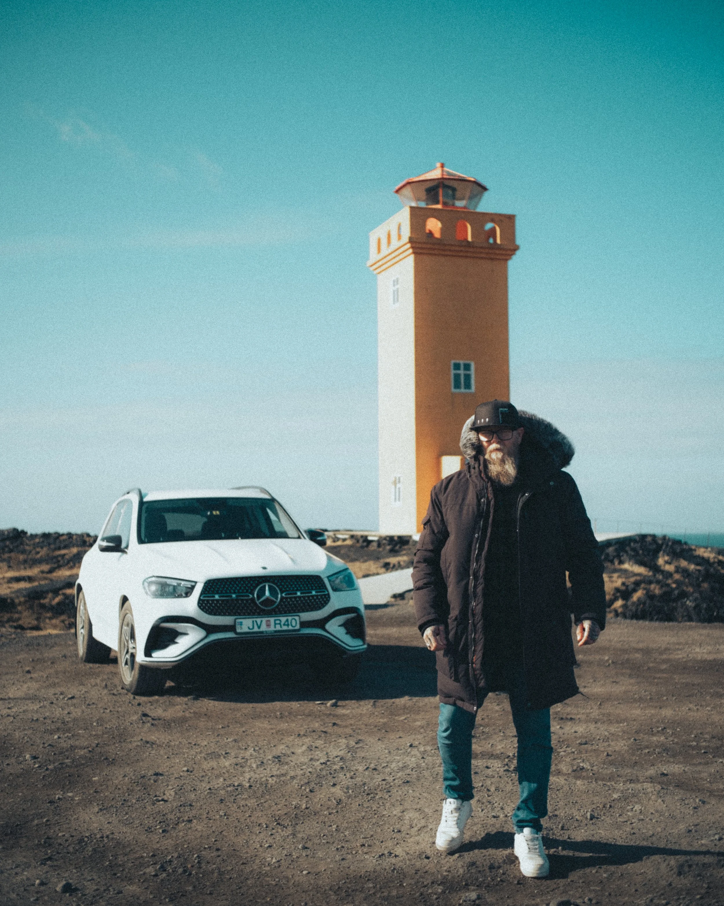 A man with a beard and glasses walking on a dirt path with a white Mercedes-Benz car behind him. In the background, there is a lighthouse and clear blue sky.