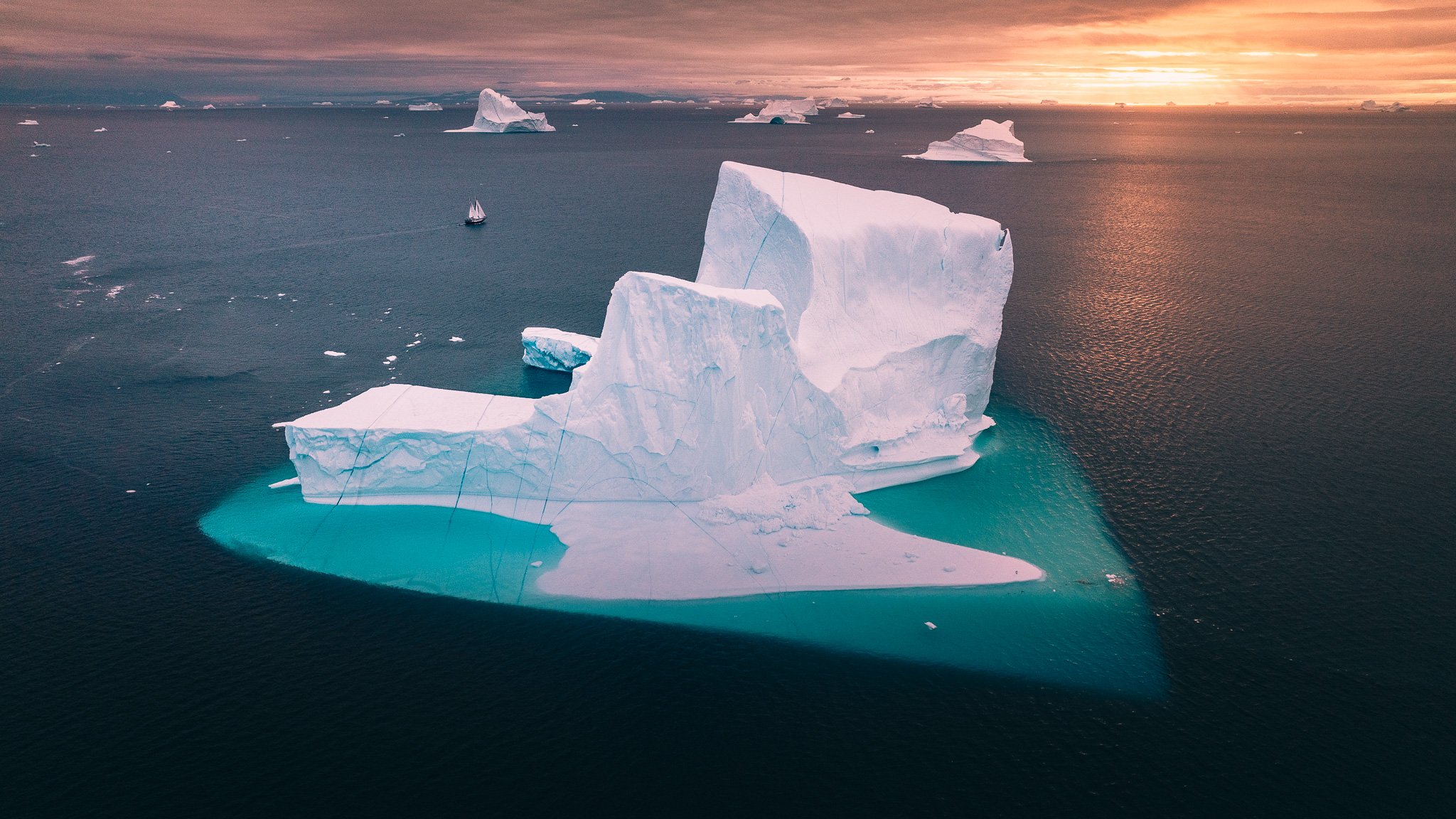 A large iceberg floating in the ocean with a ship nearby and a colorful sunset in the background.