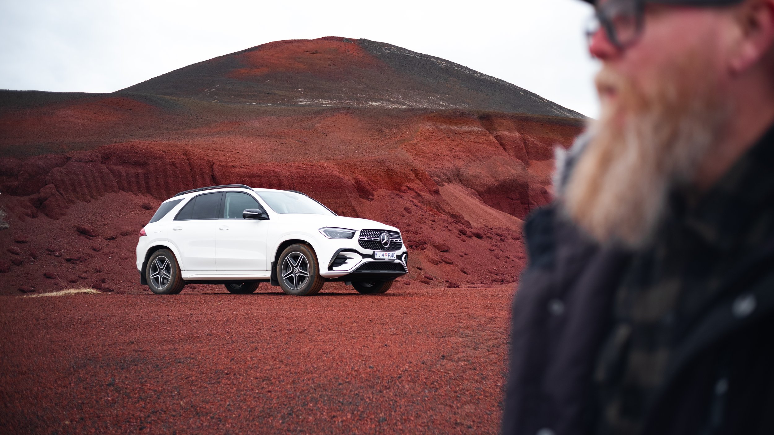 A white luxury SUV parked on a red volcanic landscape with a man in the foreground blurred out