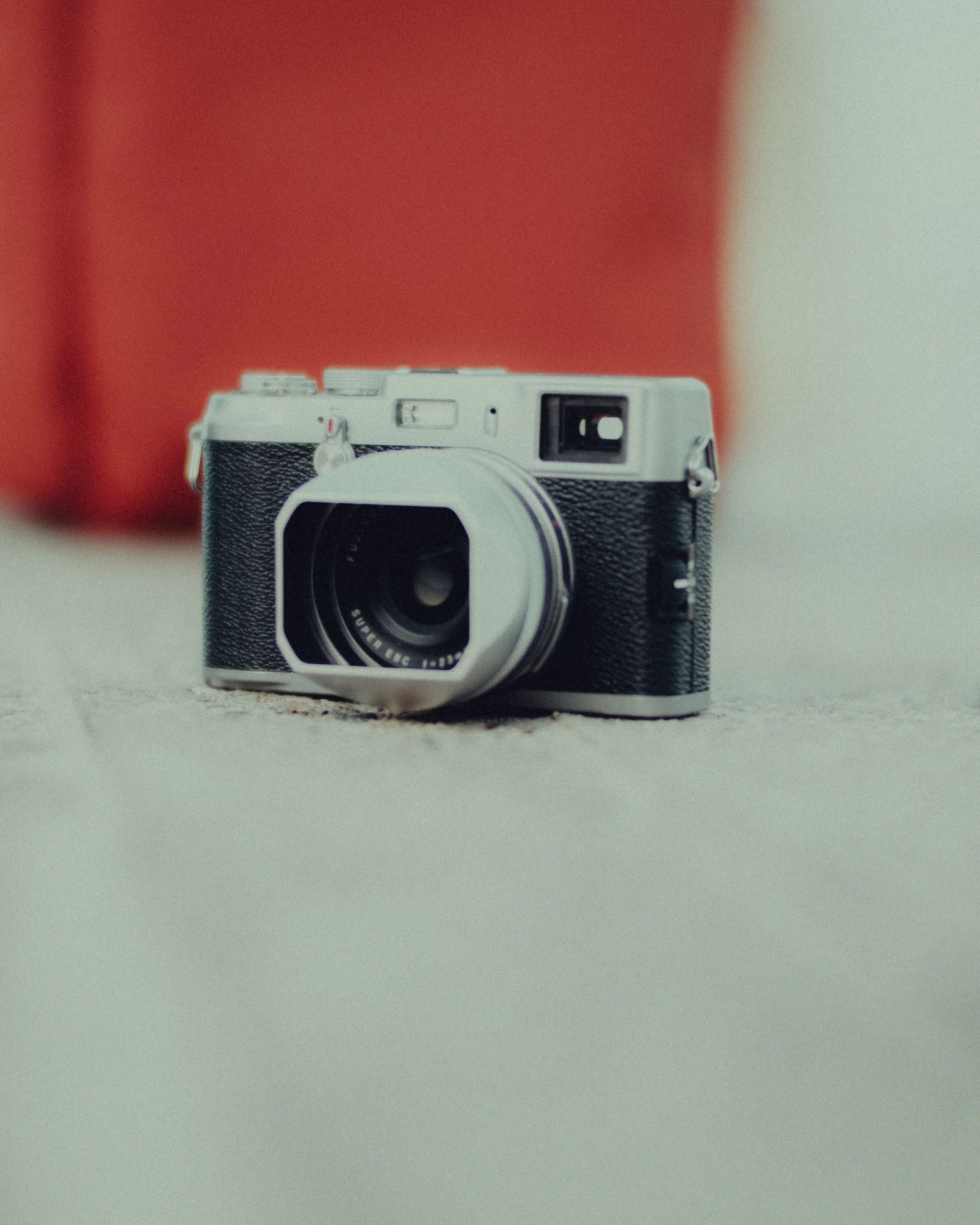 A vintage black and silver camera with a large lens sitting on a beige surface with a blurred red background.