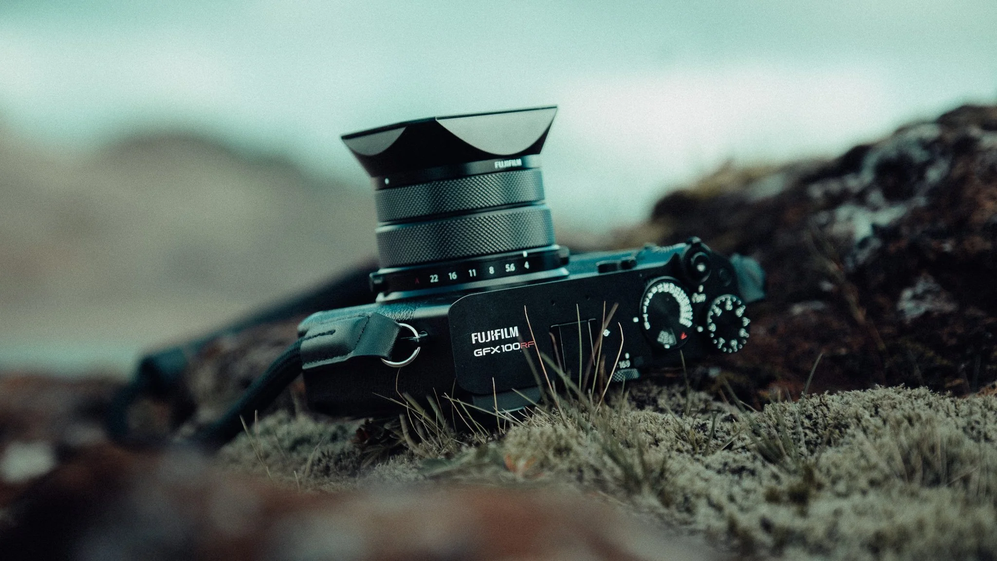 A Fujifilm camera with a lens and lens hood resting on moss and grass on a natural surface outdoors.