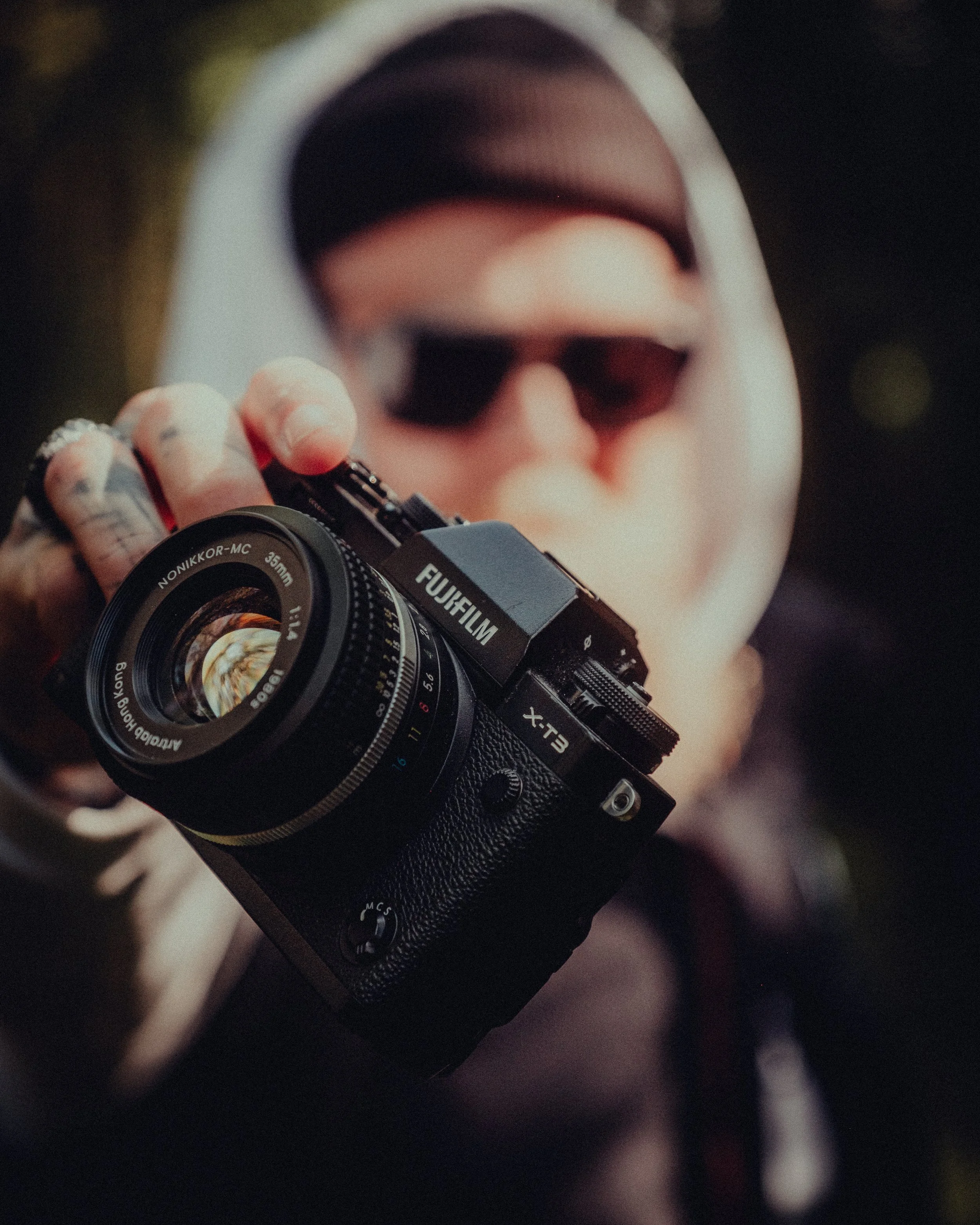 Person holding a Fujifilm camera with a 35mm lens, wearing a hoodie and sunglasses, outdoors in natural light.