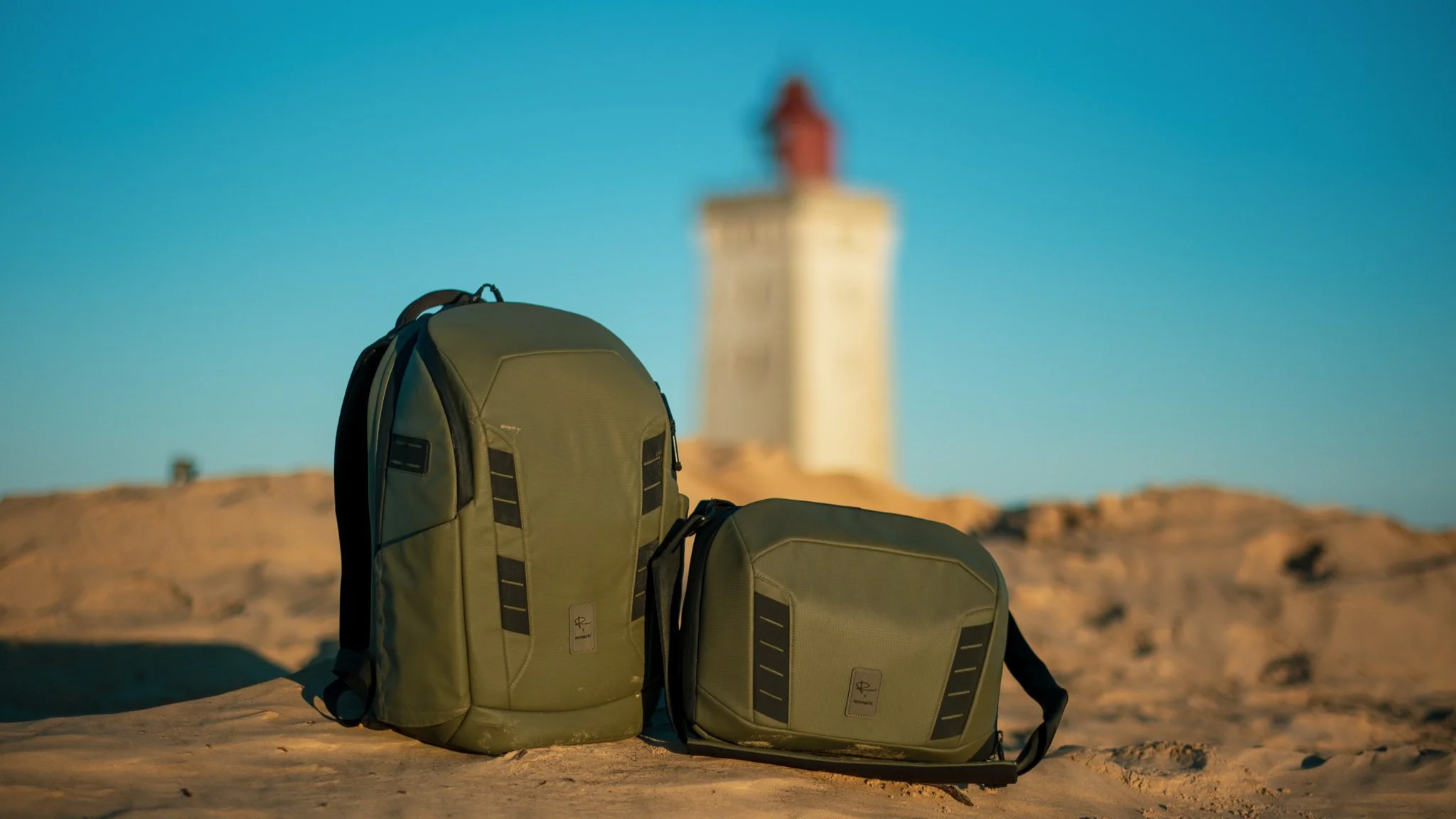 A green backpack and a green shoulder bag on sandy ground with a blurred lighthouse in the background under a clear blue sky.