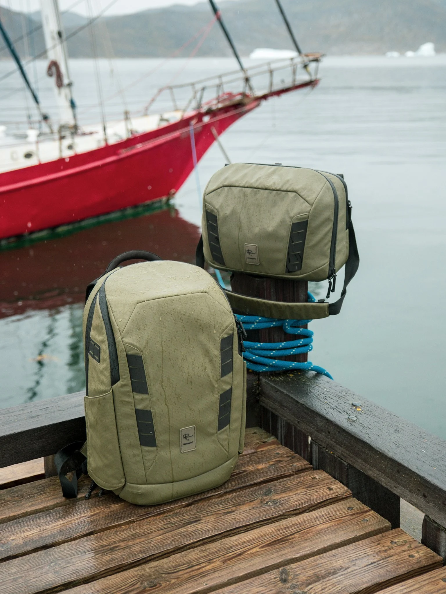 Two green backpacks resting on a wooden dock beside a body of water, with a red sailboat in the background.
