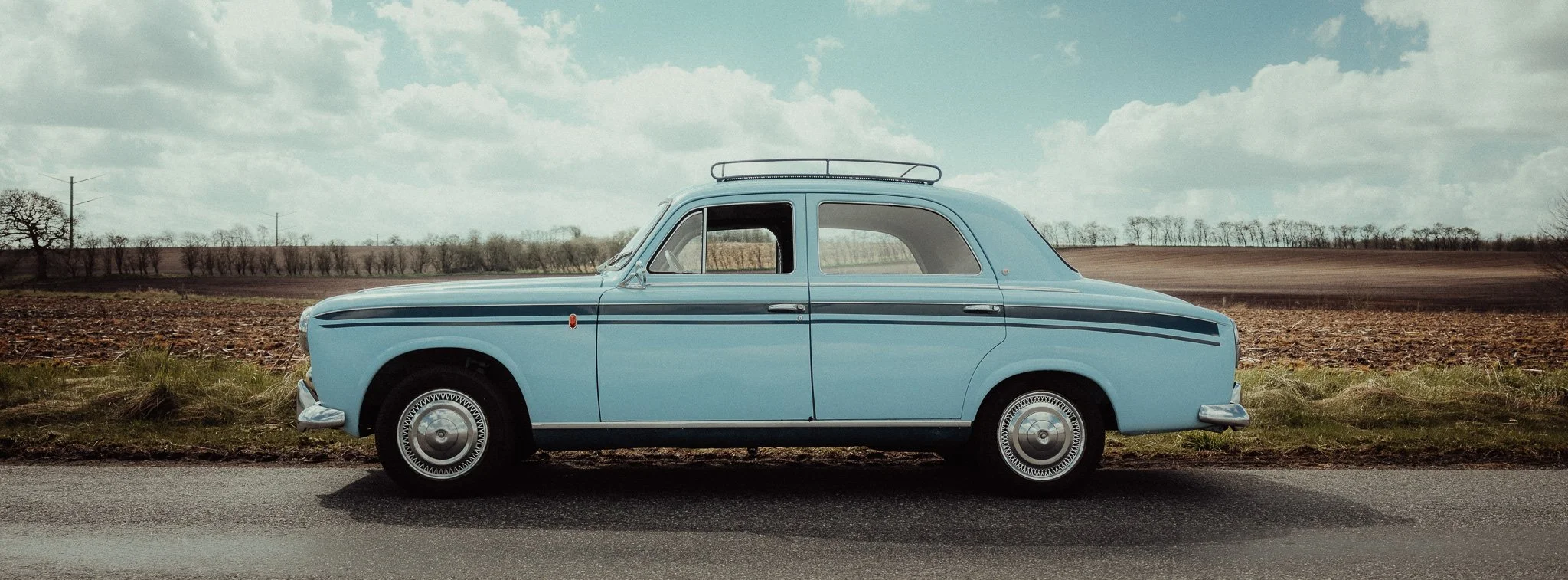 A vintage light blue car parked on the side of a rural road, with fields and a cloudy sky in the background.