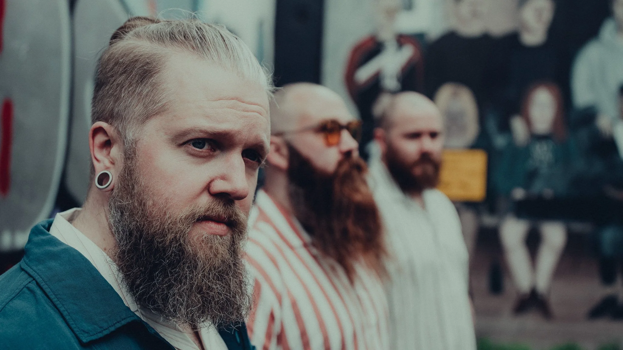 A group of men sitting in a row, with the focus on a bearded man with blond hair and gauge earrings in the foreground. The men have various facial features and hairstyles, and are wearing casual clothing, with some wearing sunglasses.