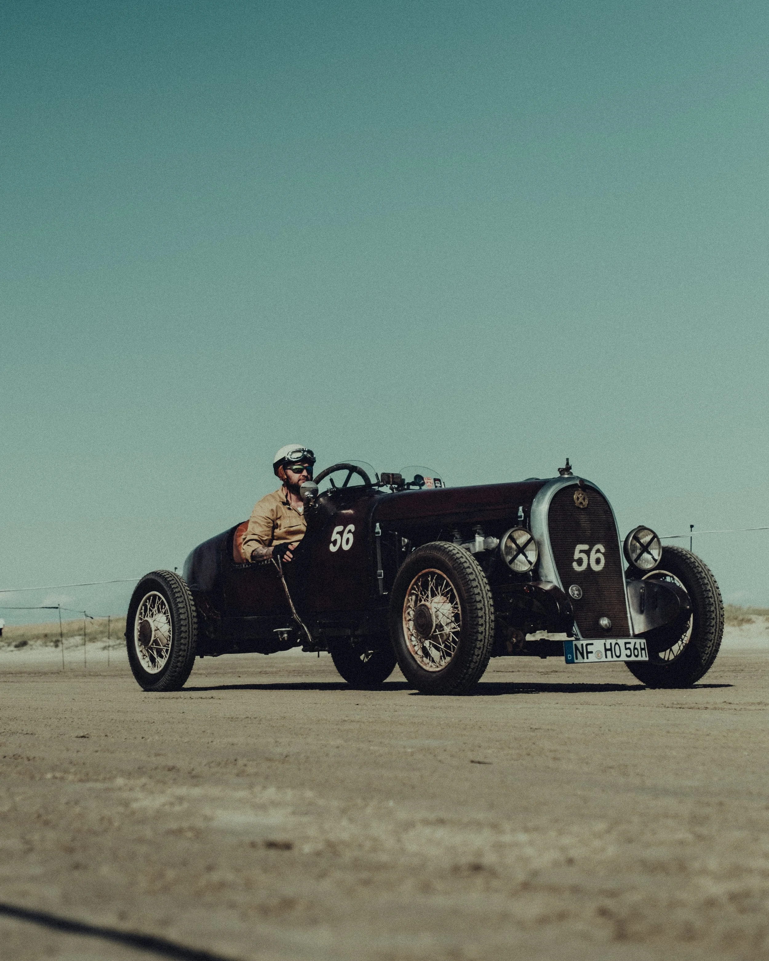 A vintage black race car with the number 56, driven by a man wearing a helmet and goggles, on a dirt track in a desert landscape.