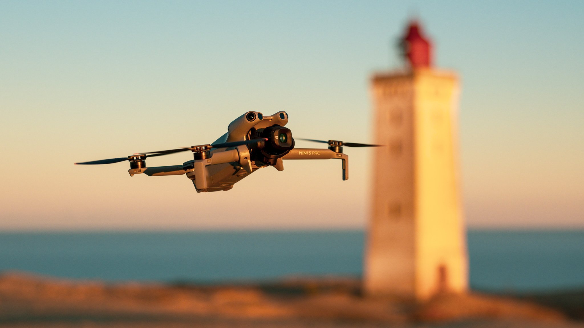 A drone flying near a lighthouse during sunset or sunrise with a blurred horizon in the background.