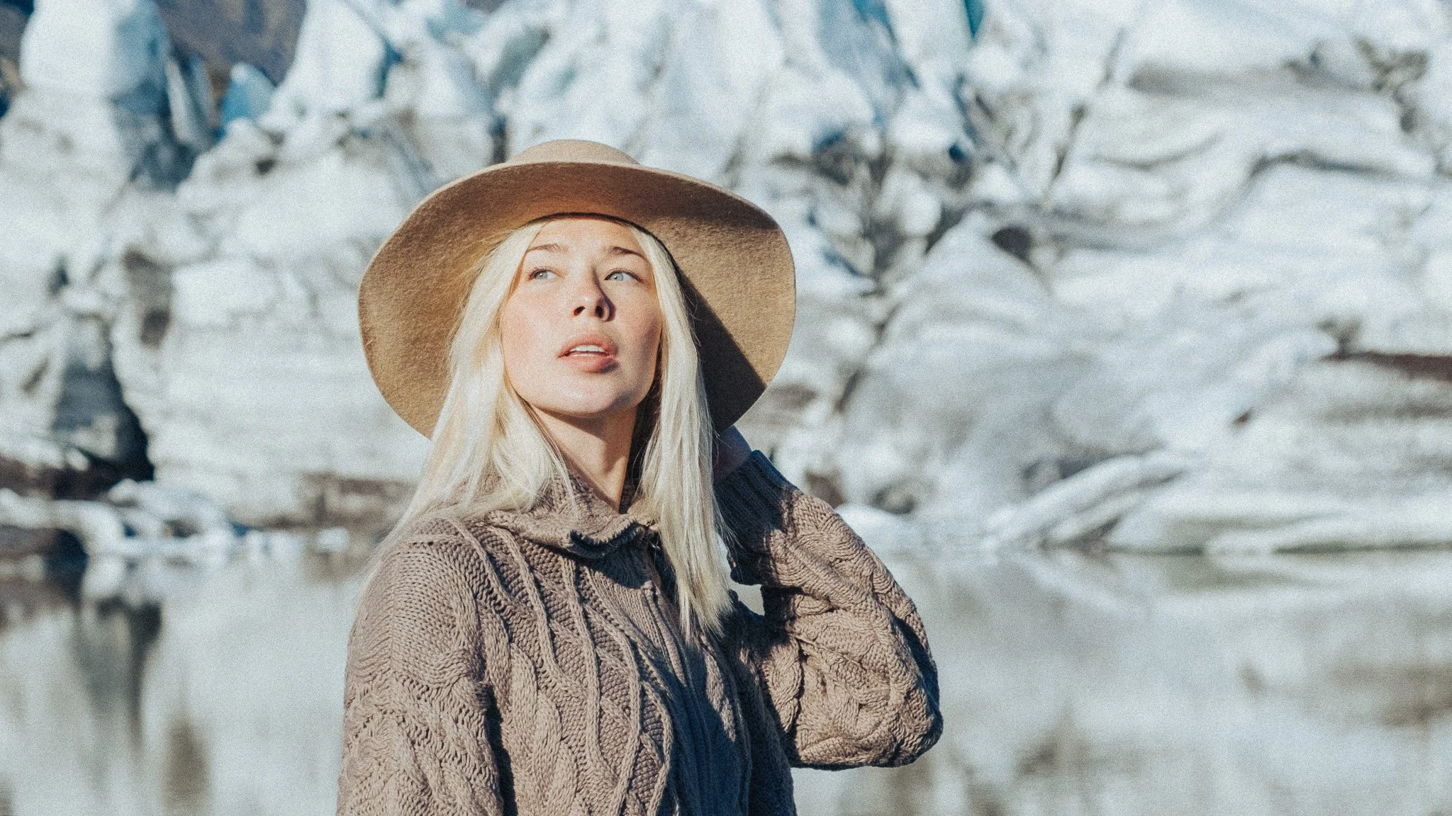 A young woman with long blonde hair wearing a large beige sun hat and a knitted brown sweater, standing outdoors with snow-covered trees in the background.