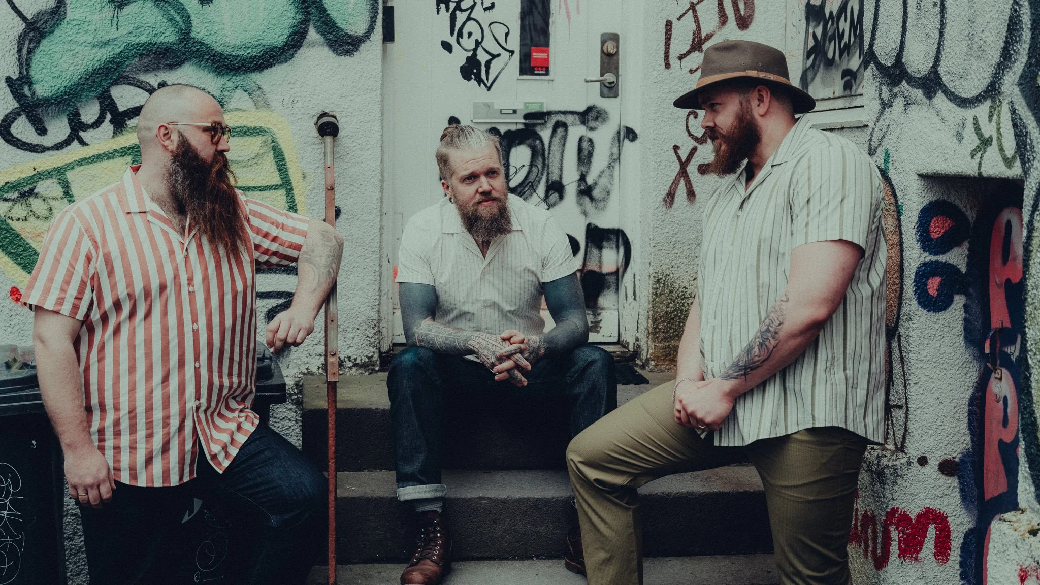 Three men with tattoos and beards are standing and sitting against a graffiti-covered wall, having a conversation outdoors.