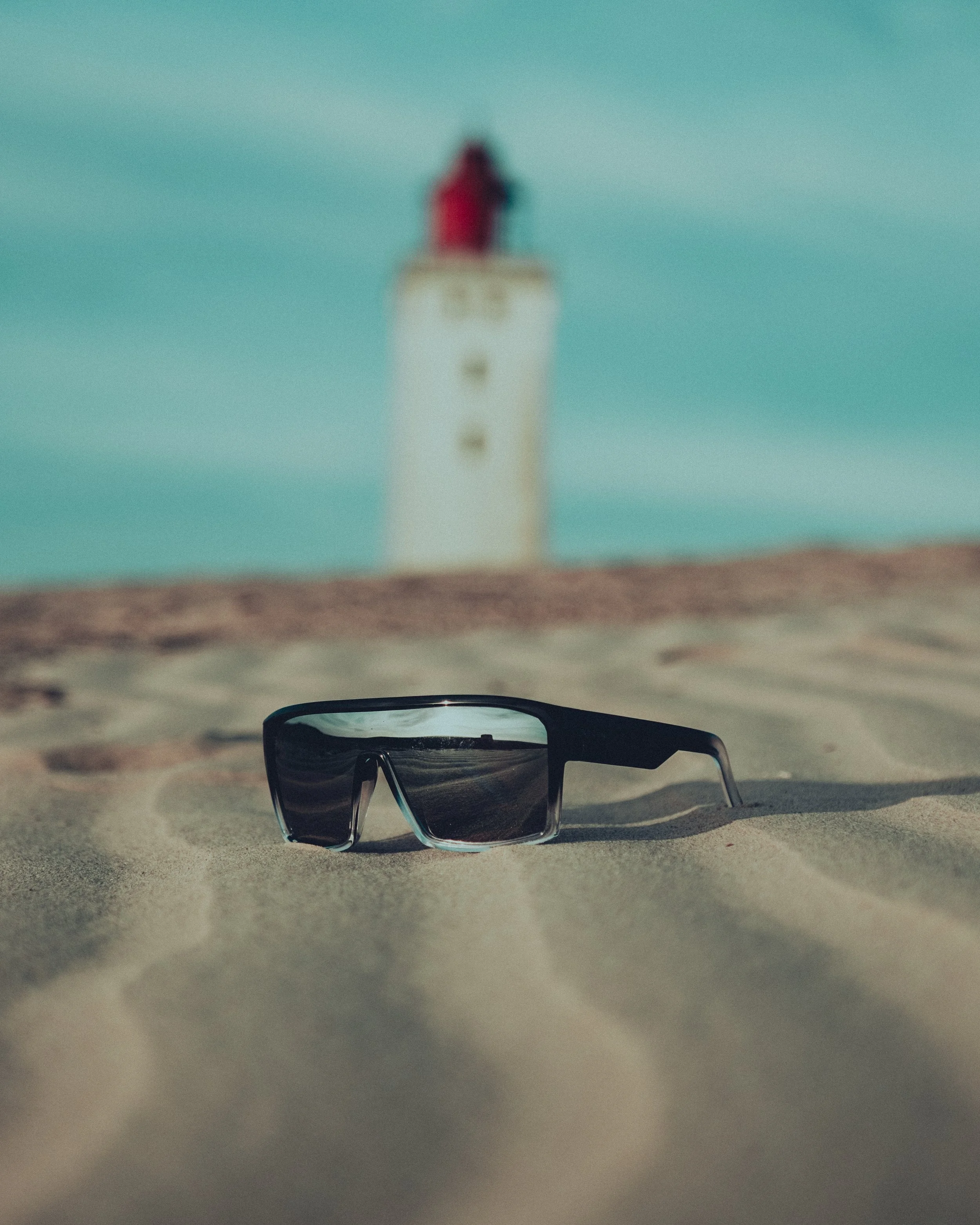 Sunglasses on sandy ground with a blurred lighthouse in the background.
