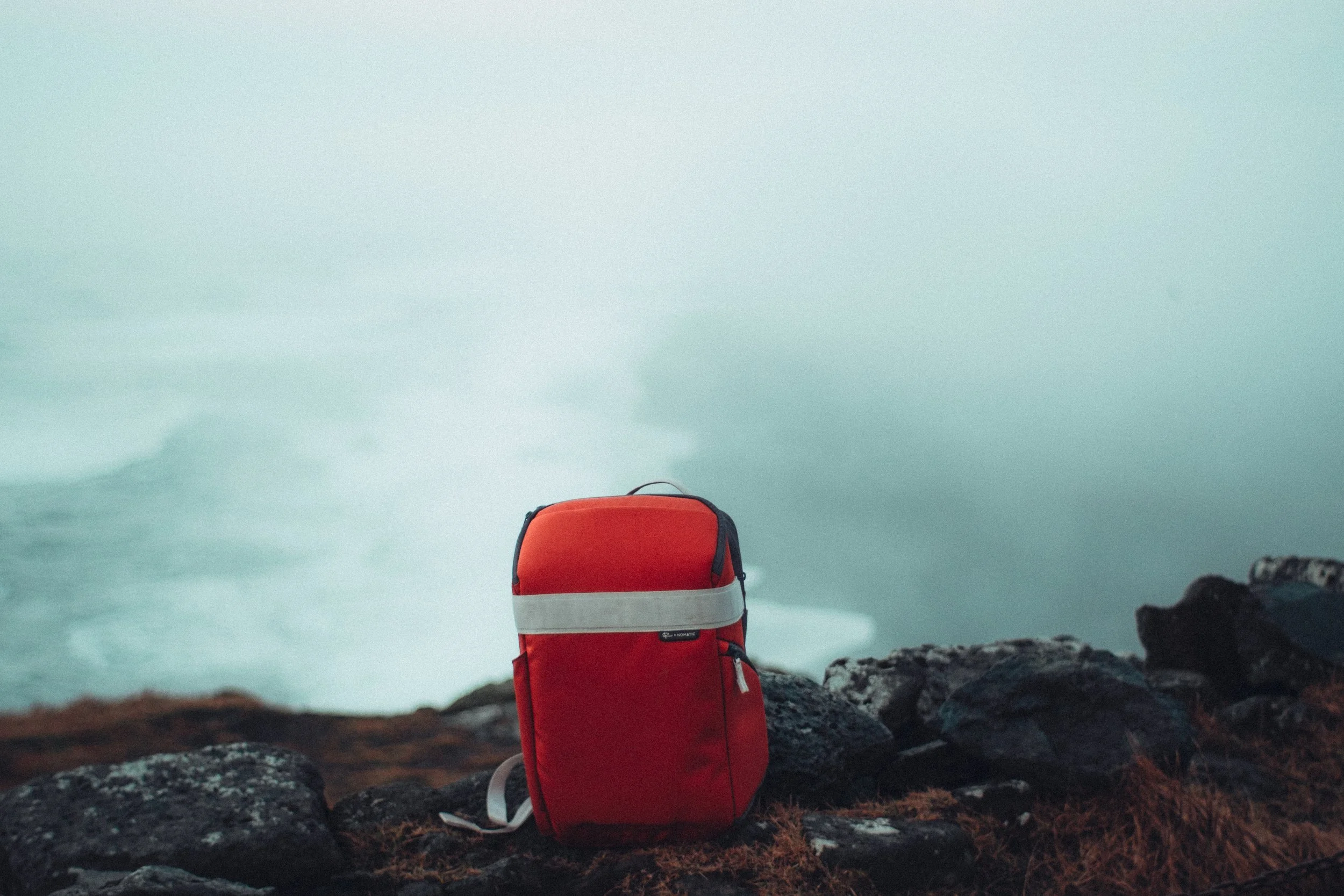 A red backpack resting on rocks near a foggy body of water or ocean in the distance.