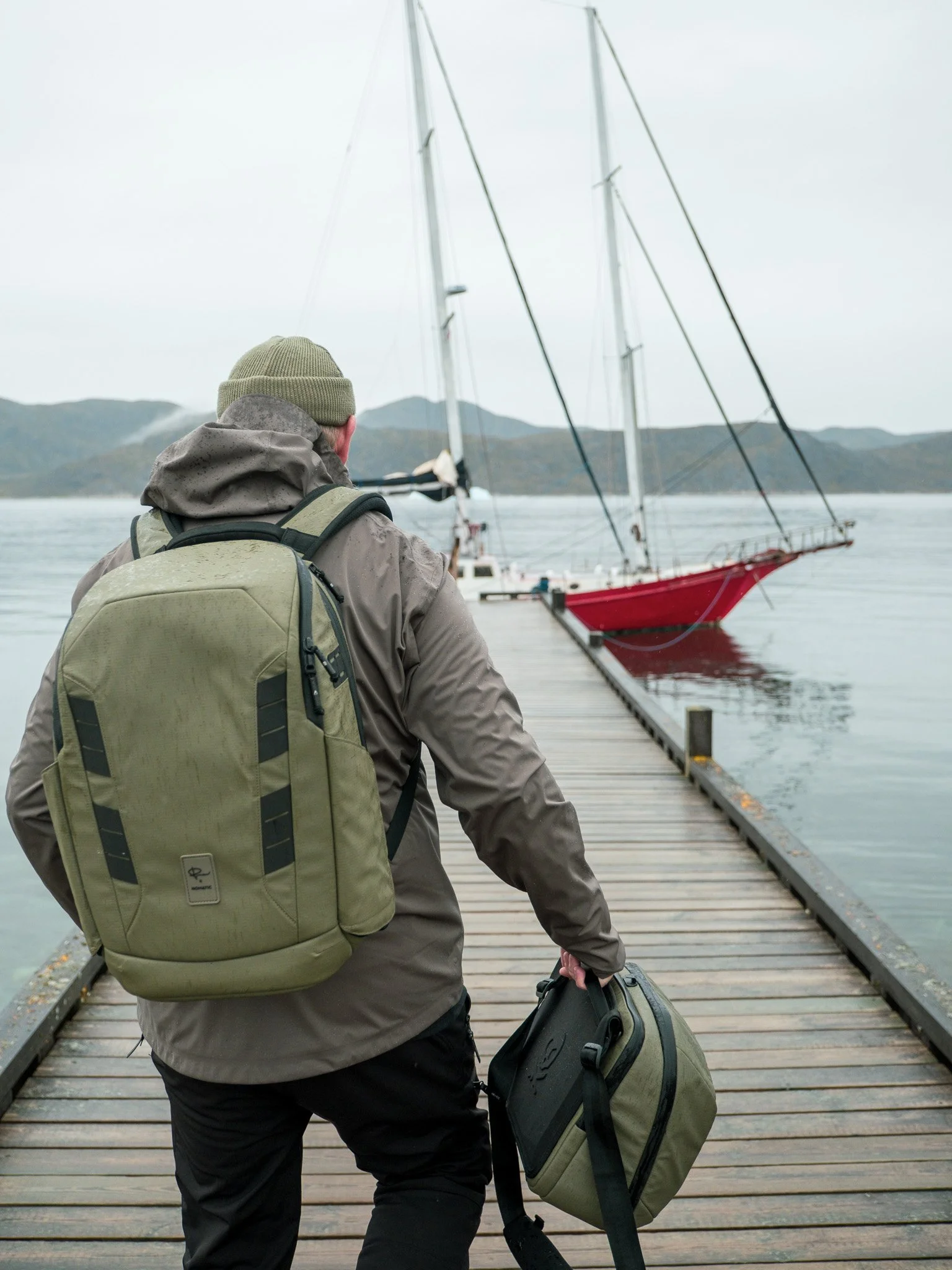 Person in outdoor gear walking on a dock carrying a backpack and a bag, with a sailboat moored at the dock on a cloudy day.
