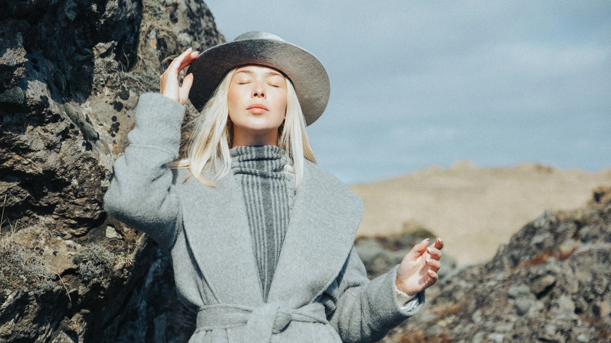 A woman with blonde hair and closed eyes, wearing a gray coat and a plaid scarf, stands outdoors with a rocky landscape in the background, adjusting a wide-brimmed hat on her head.