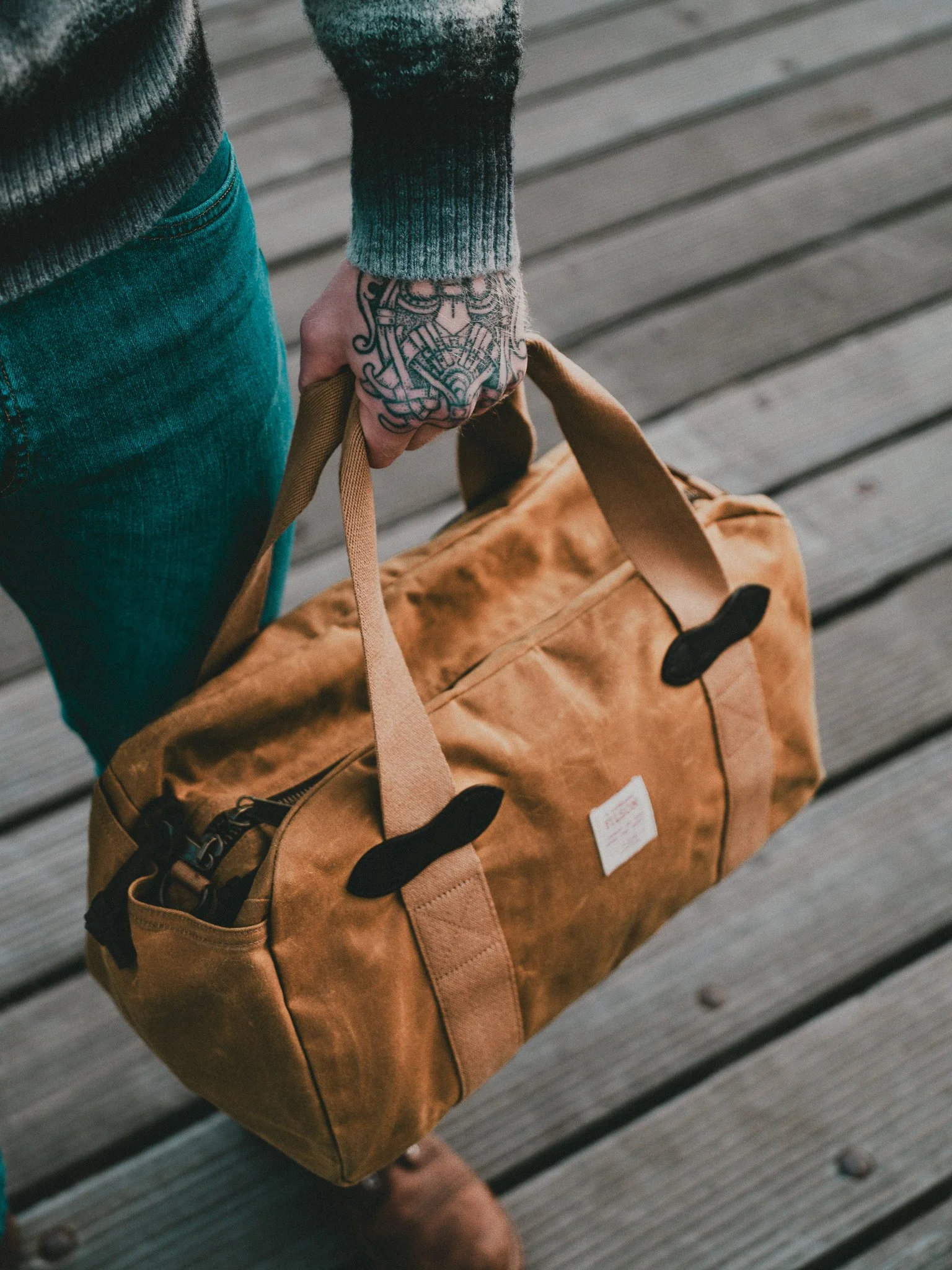 Close-up of a person's hand holding a brown duffel bag with a patch pocket and black accents. The person is wearing a dark sweater with a grayish-green cuff and blue jeans. The background shows a wooden deck with parallel planks.