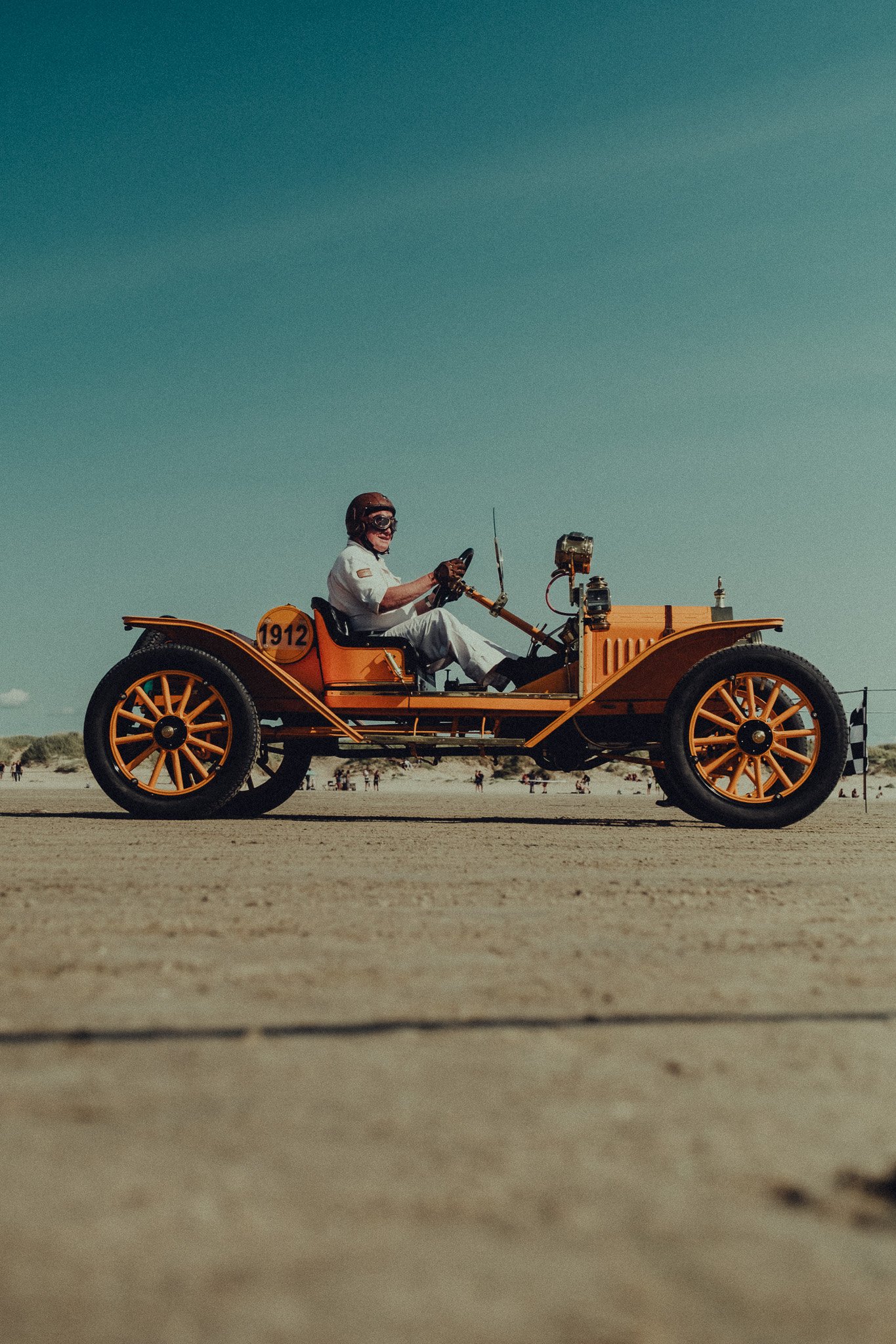 A person dressed in a white outfit, wearing goggles and a helmet, sitting in an orange vintage-style car with the number 1912 on its side. The car is on a sandy beach with many people in the background.