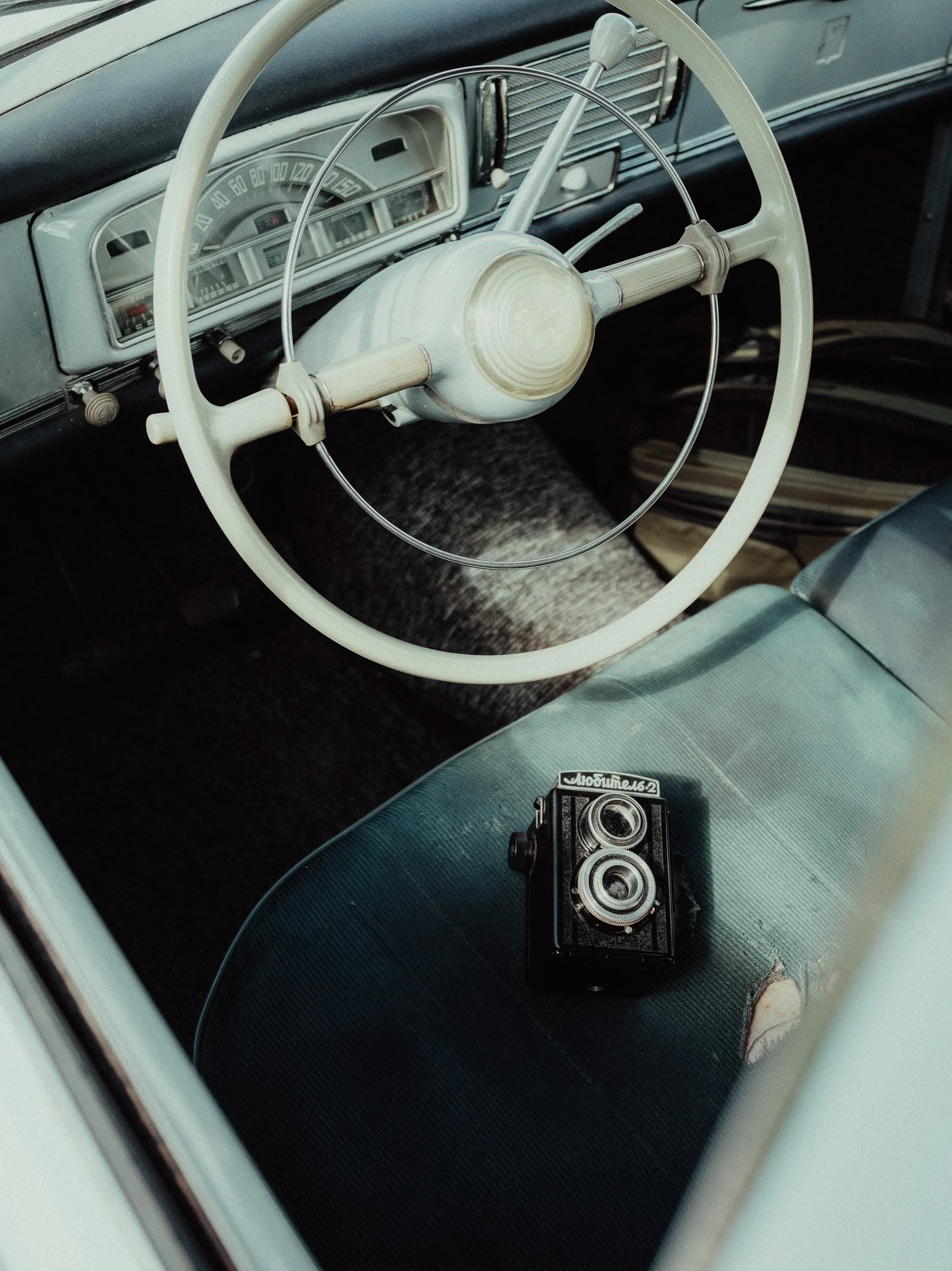 Inside an older car, showing a white steering wheel, a speedometer, and a vintage camera on the seat.