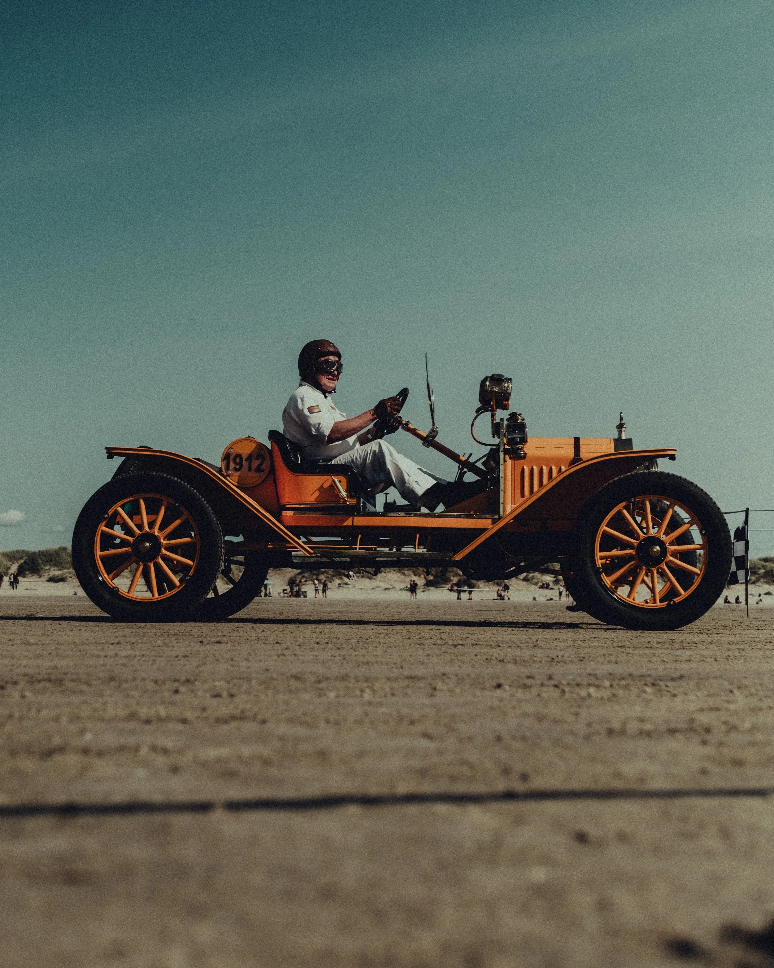 A person driving a vintage orange race car on a beach with a clear sky in the background.