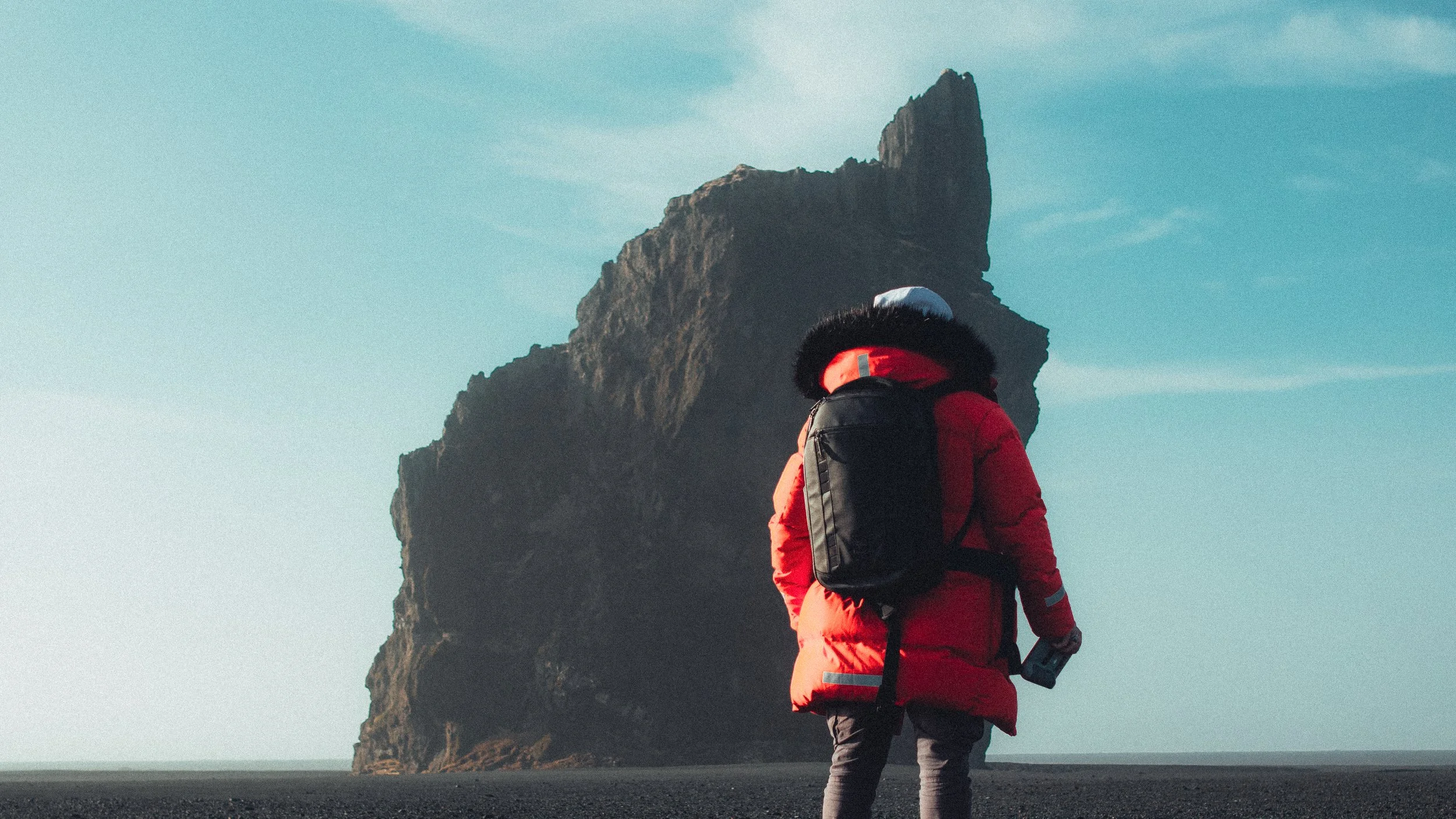 Person in red winter coat with a backpack looking at a large rocky mountain on a dark beach under a cloudy sky.