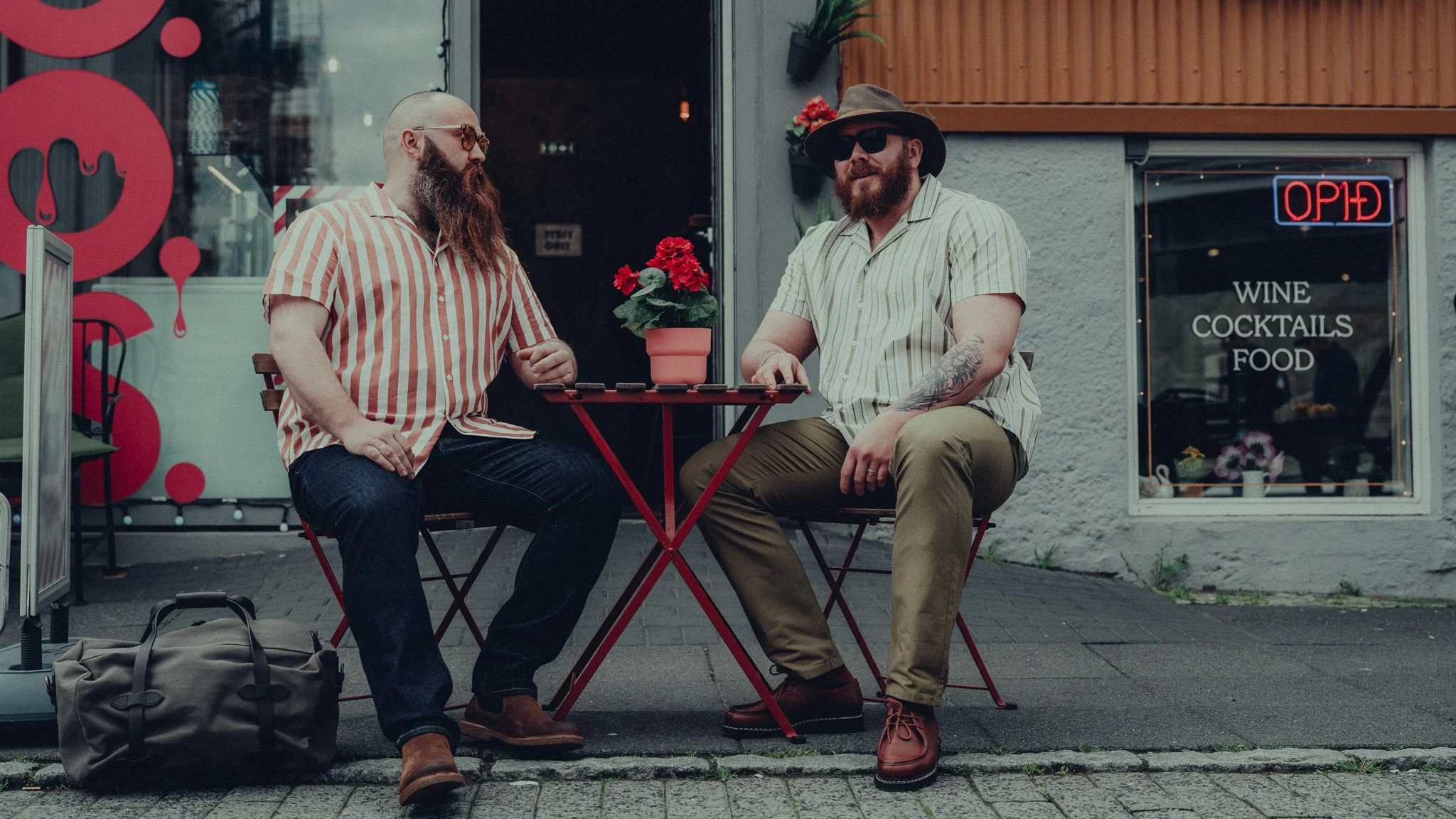 Two men sitting at a small outdoor table outside a cafe, engaging in conversation. One has a long red beard and glasses, wearing a striped short-sleeve shirt and jeans. The other has a beard and is wearing a wide-brimmed hat, sunglasses, a striped sh