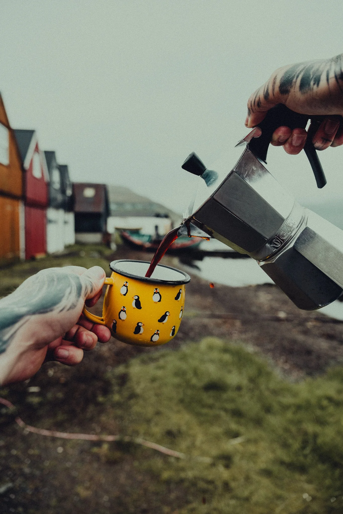 Person pouring coffee into a penguin-themed mug outdoors, with colorful barn-like buildings in the background.