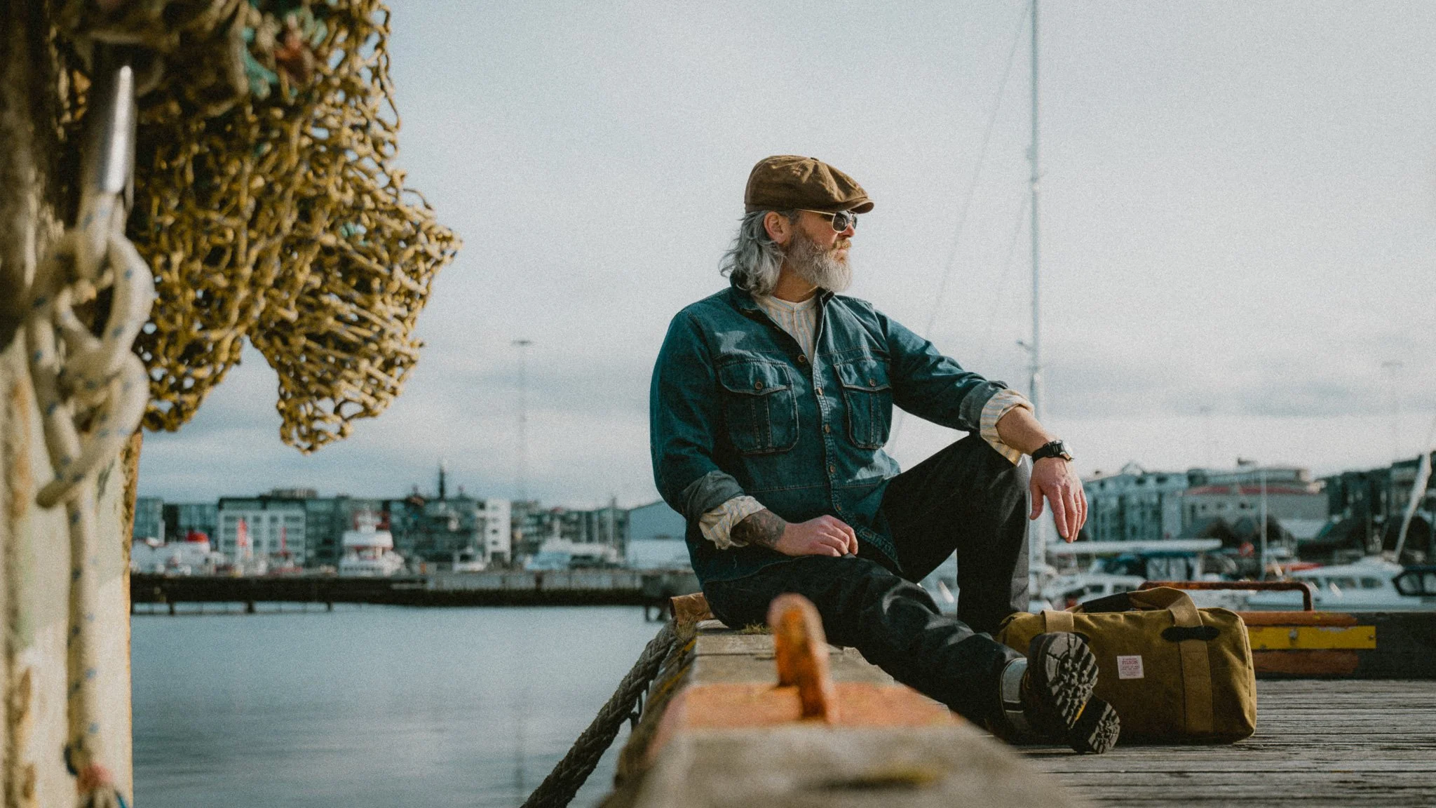 A man with long gray hair, beard, glasses, wearing a brown cap, denim jacket, and striped shirt, sitting on a dock by the water with boats and city buildings in the background, during daytime.