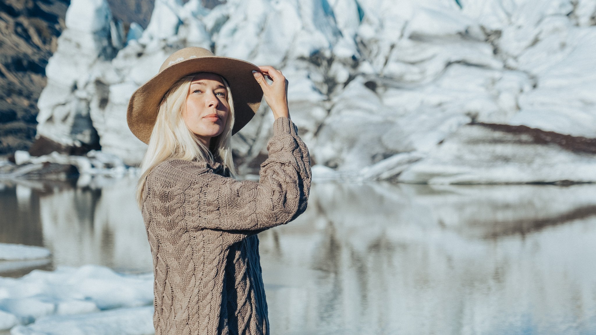 A woman with long blonde hair wearing a wide-brimmed hat and a brown knit sweater stands near a body of water in a snowy landscape, with ice and snow-covered rocks in the background.