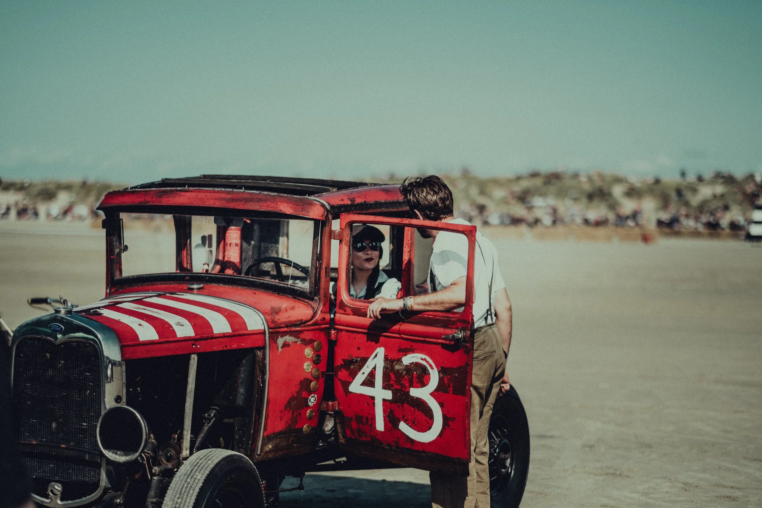 Two people conversing near a vintage red race car with the number 43, parked on a flat, open area with a distant crowd in the background.
