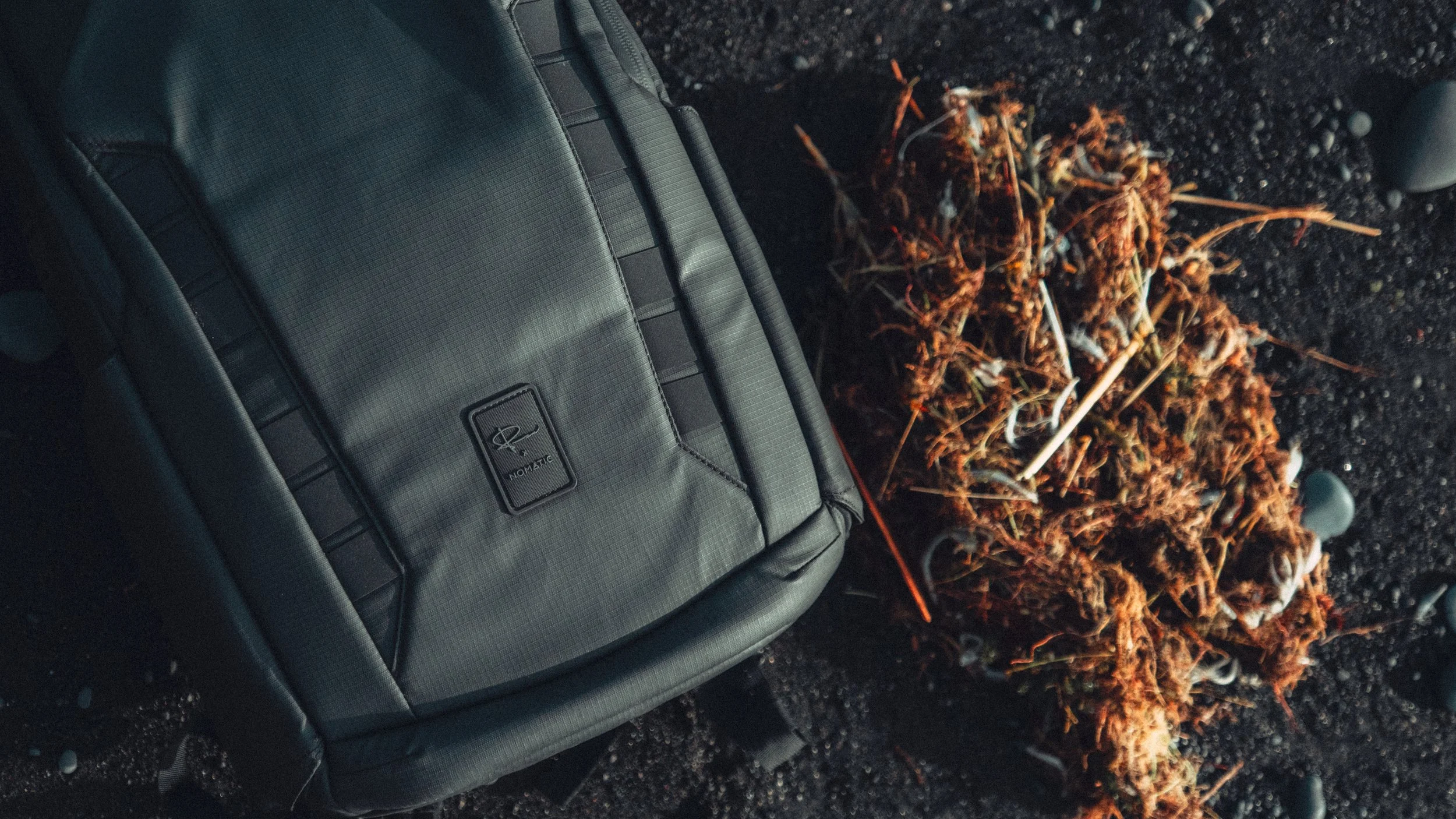 A gray Nomatic backpack on black volcanic sand next to a pile of burnt, dried wood and twigs.