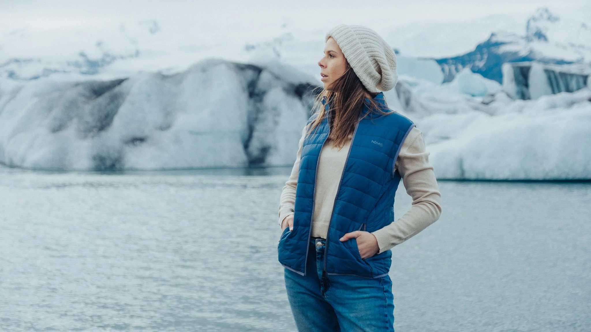 A woman standing near a body of water with glaciers in the background, wearing a white knit hat and a blue puffy vest.
