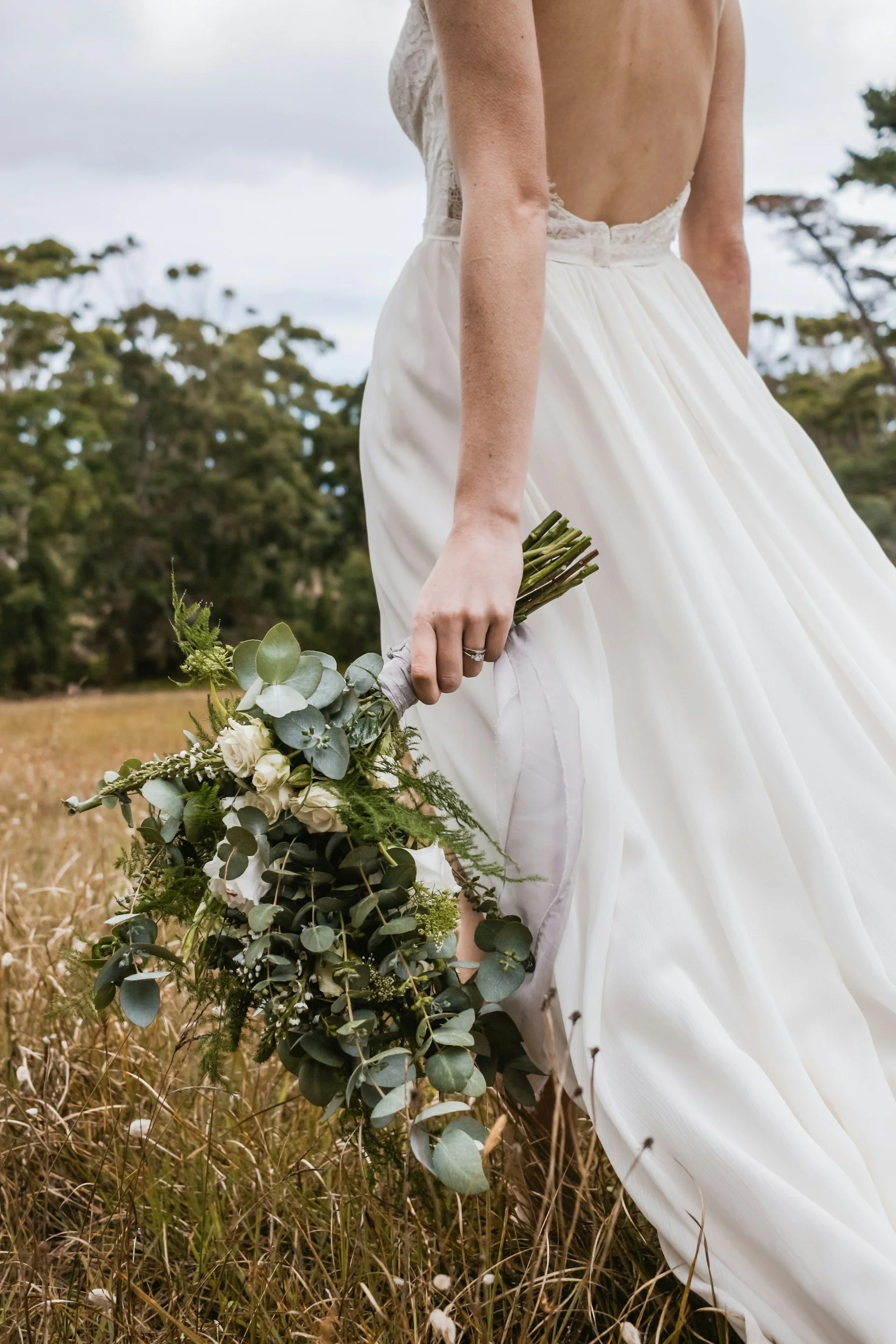 Bride holding a bouquet of white flowers and greenery in a field, wearing a white wedding dress with an open back.