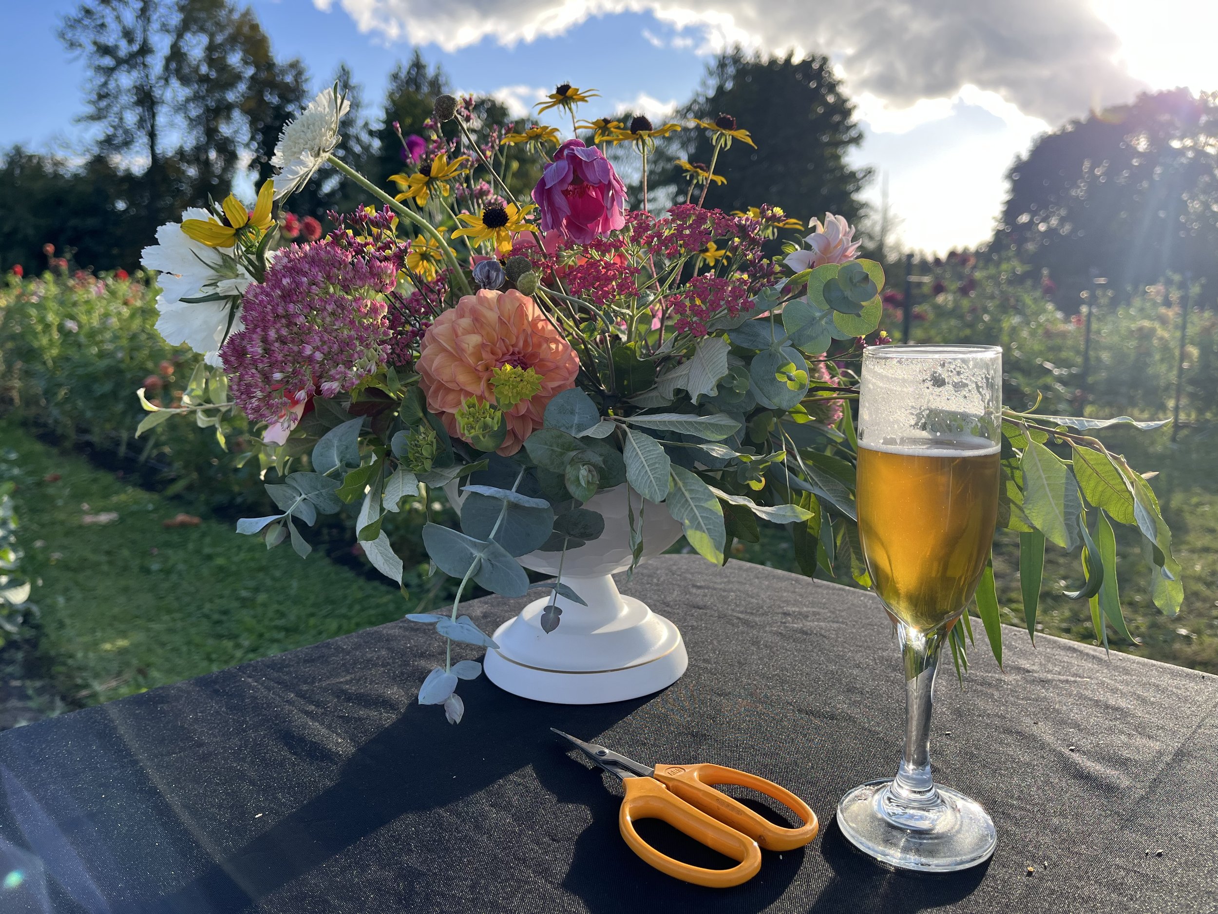 A bouquet of colorful flowers in a white vase, a pair of orange scissors, and a glass of beer on a black table outdoors during sunset.