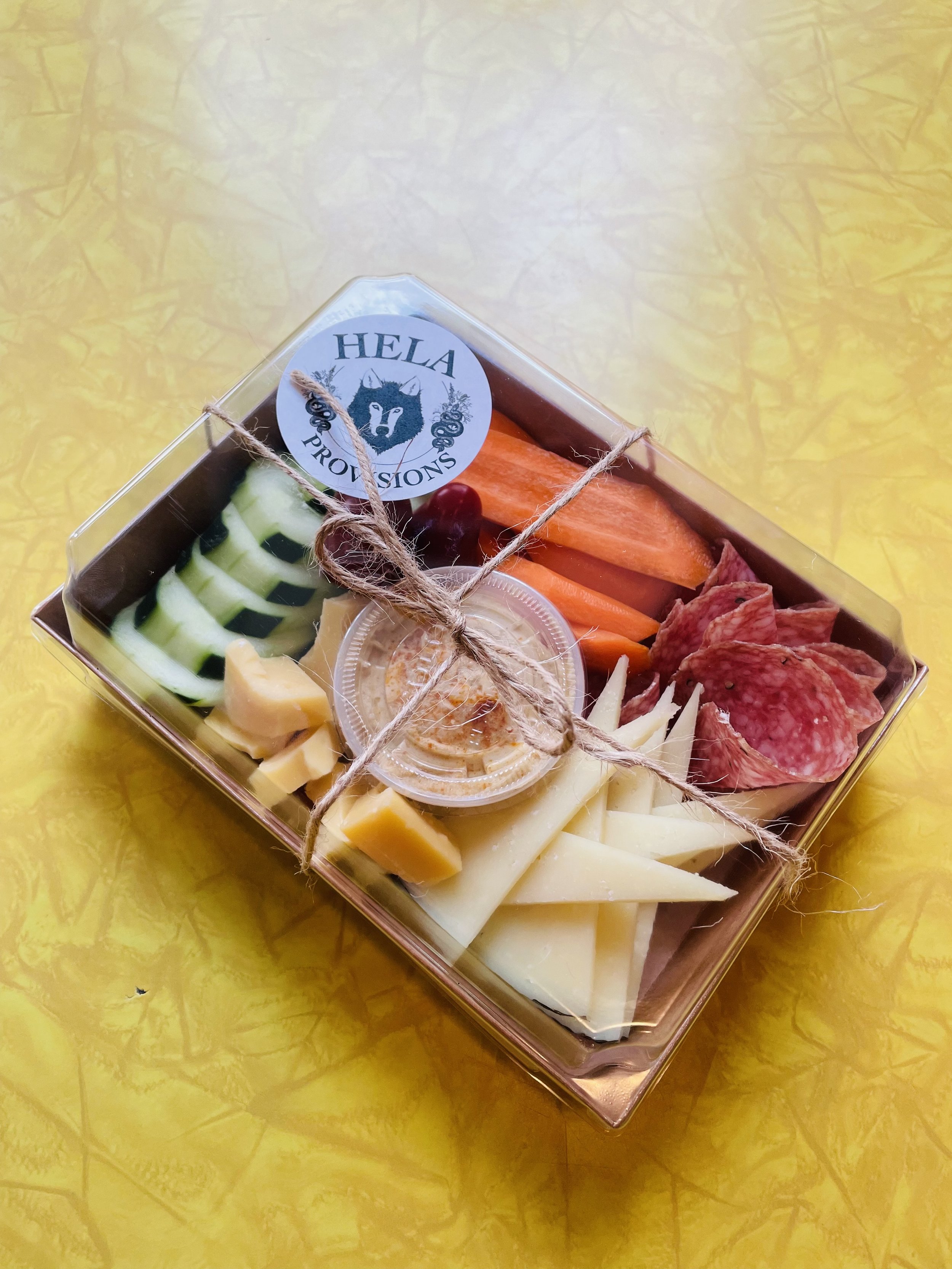 A charcuterie box with sliced cucumbers, cheese, carrots, pepperoni, and a small container of dip, sealed with a sticker that reads 'Hela Provisions' with a wolf logo, placed on a yellow surface.