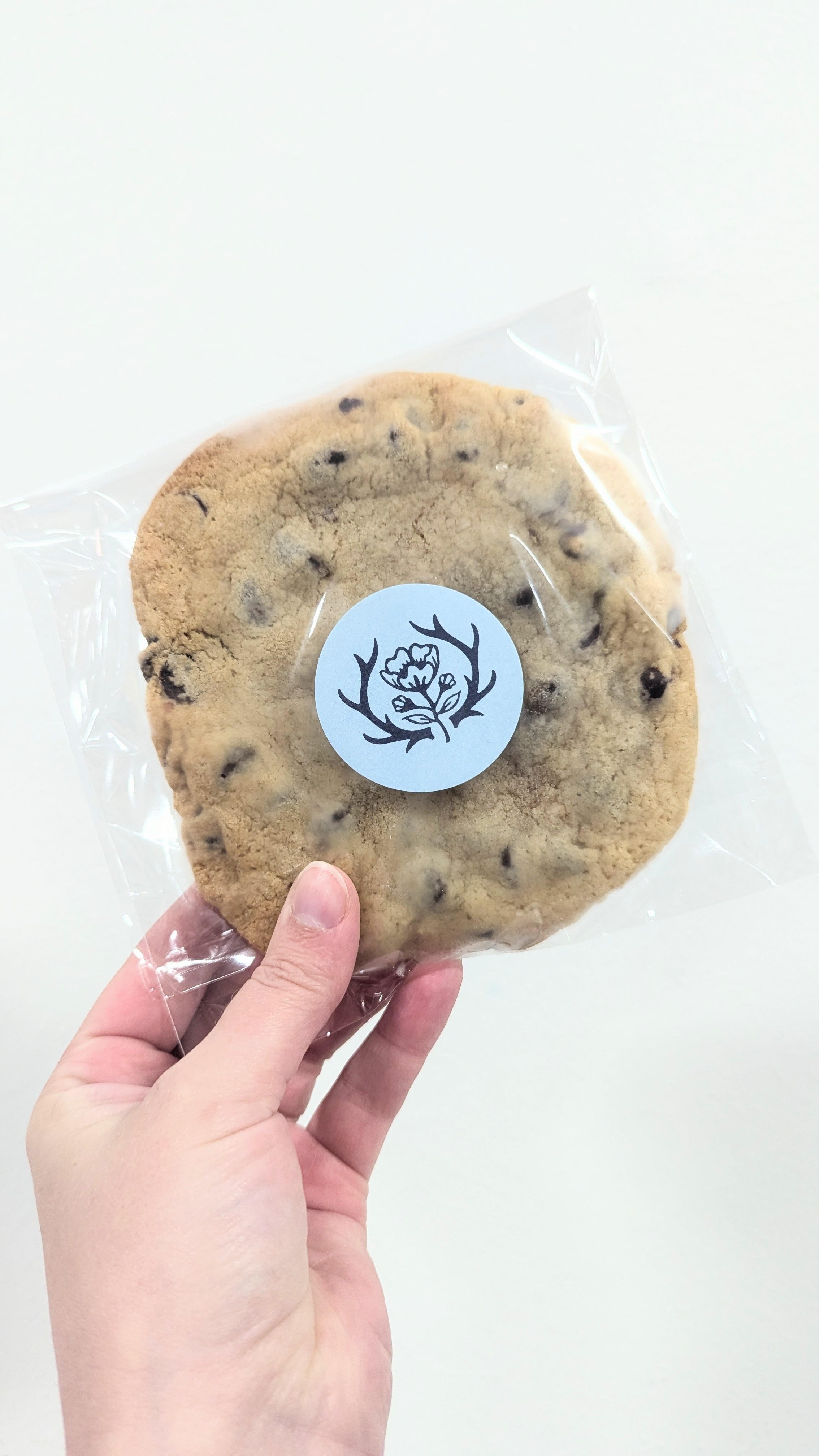A person holding a large chocolate chip cookie in a clear plastic bag with a round sticker featuring a flower and antlers logo.