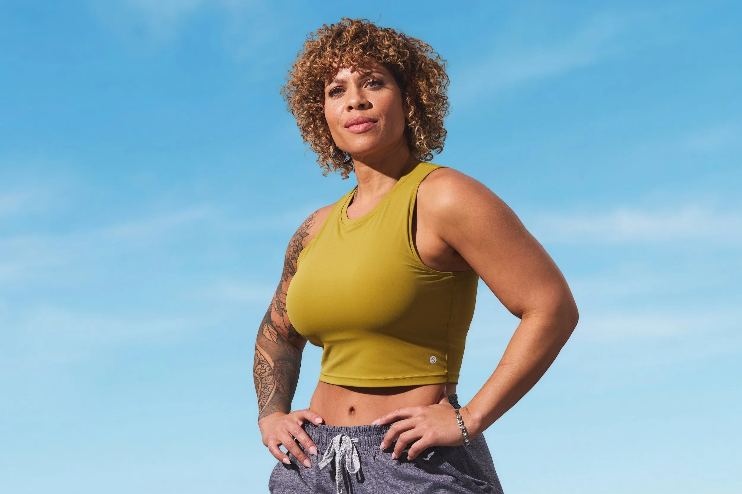 Woman with curly hair poses confidently outside, wearing a yellow crop top and gray pants, with a tattoo on her arm, against a blue sky background.