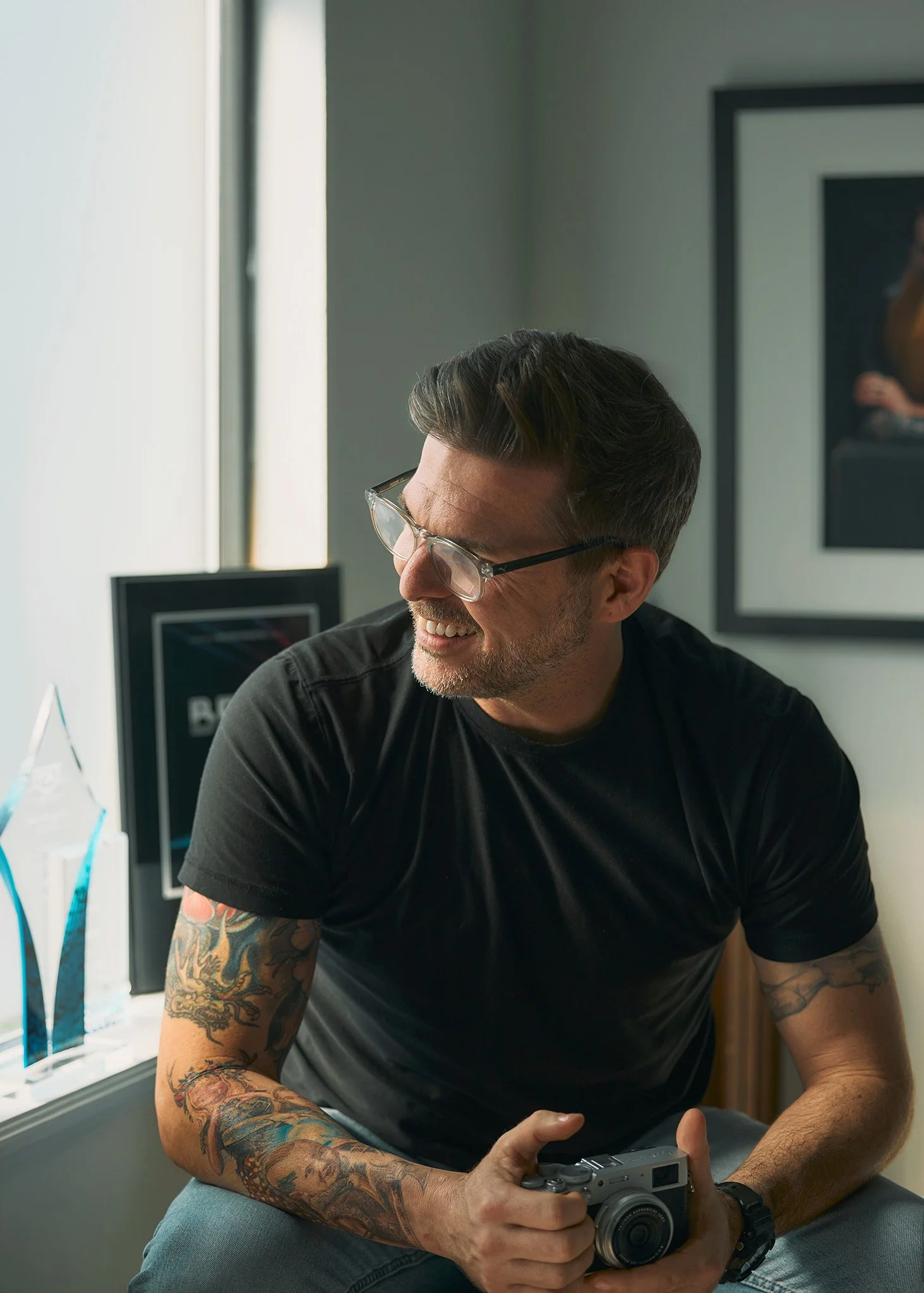 A person with tattoos sitting near a window, holding a camera and smiling, with awards and framed pictures in the background.