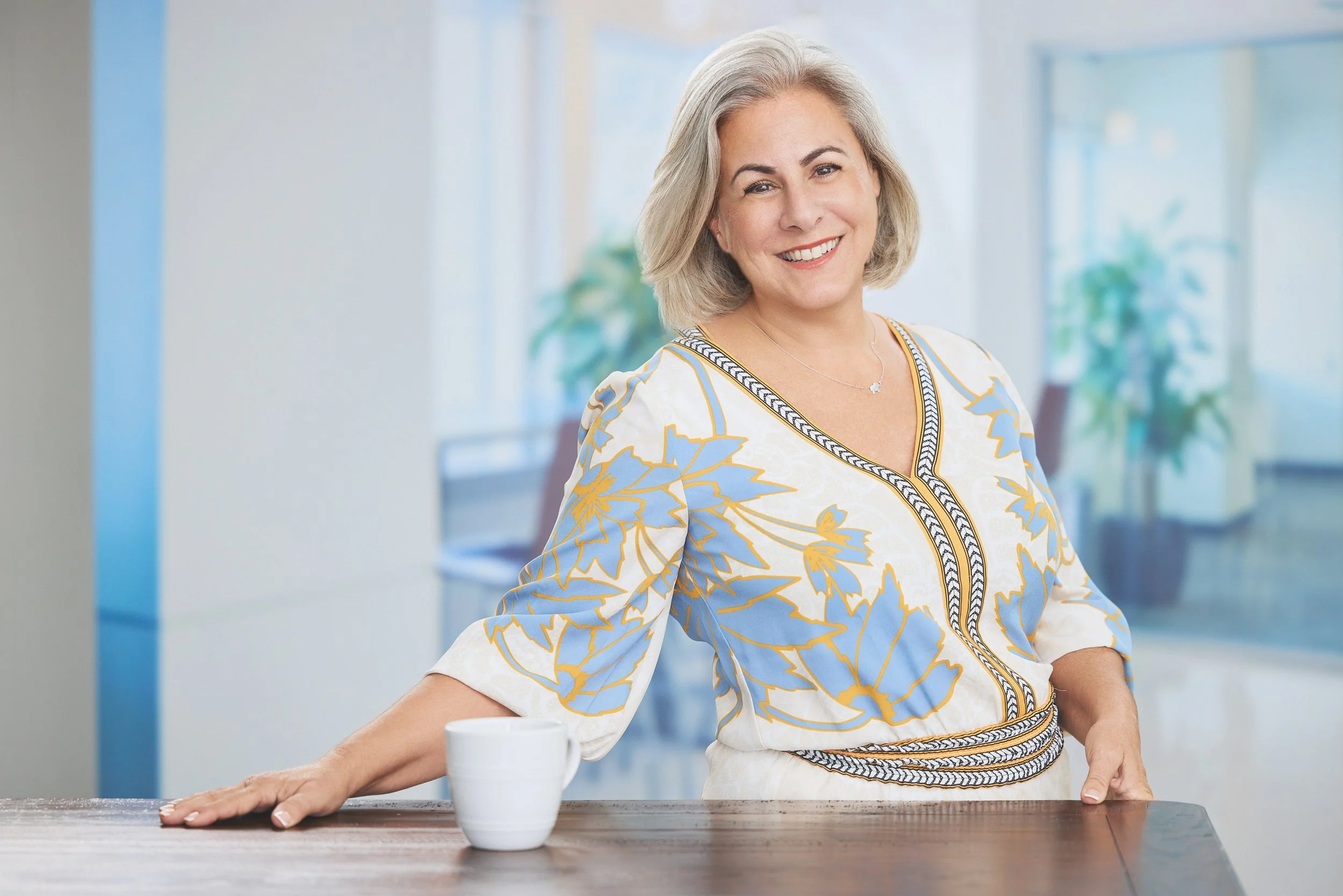 Smiling woman in a floral blouse leaning on a wooden table with a white mug in a bright office setting.