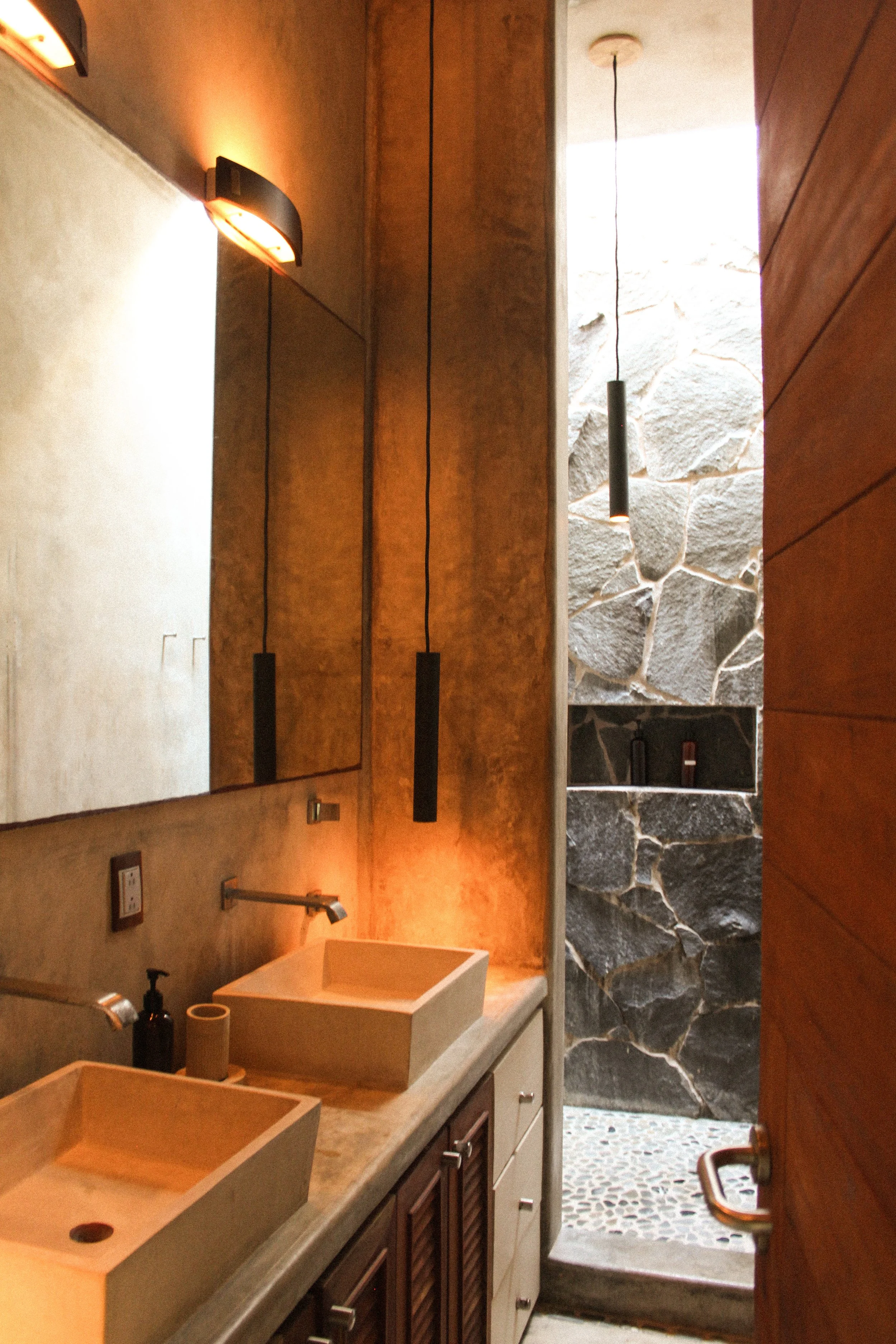 Modern bathroom with dual vessel sinks, warm lighting, mirror, and a stone shower area with a narrow window above.