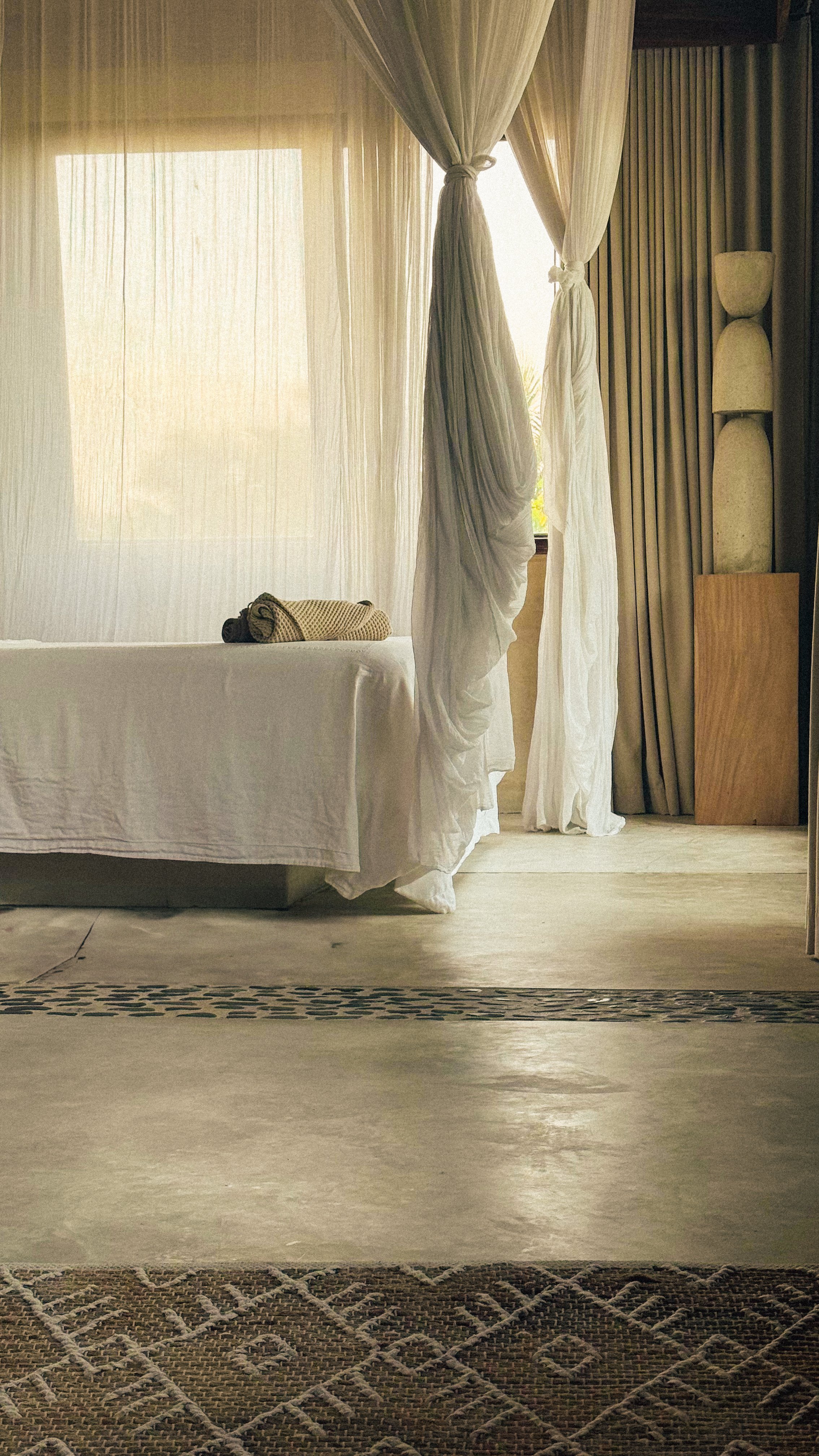 A bedroom with sheer curtains, a bed with rolled towel, and a wooden headboard. Sunlight filters through the curtains.