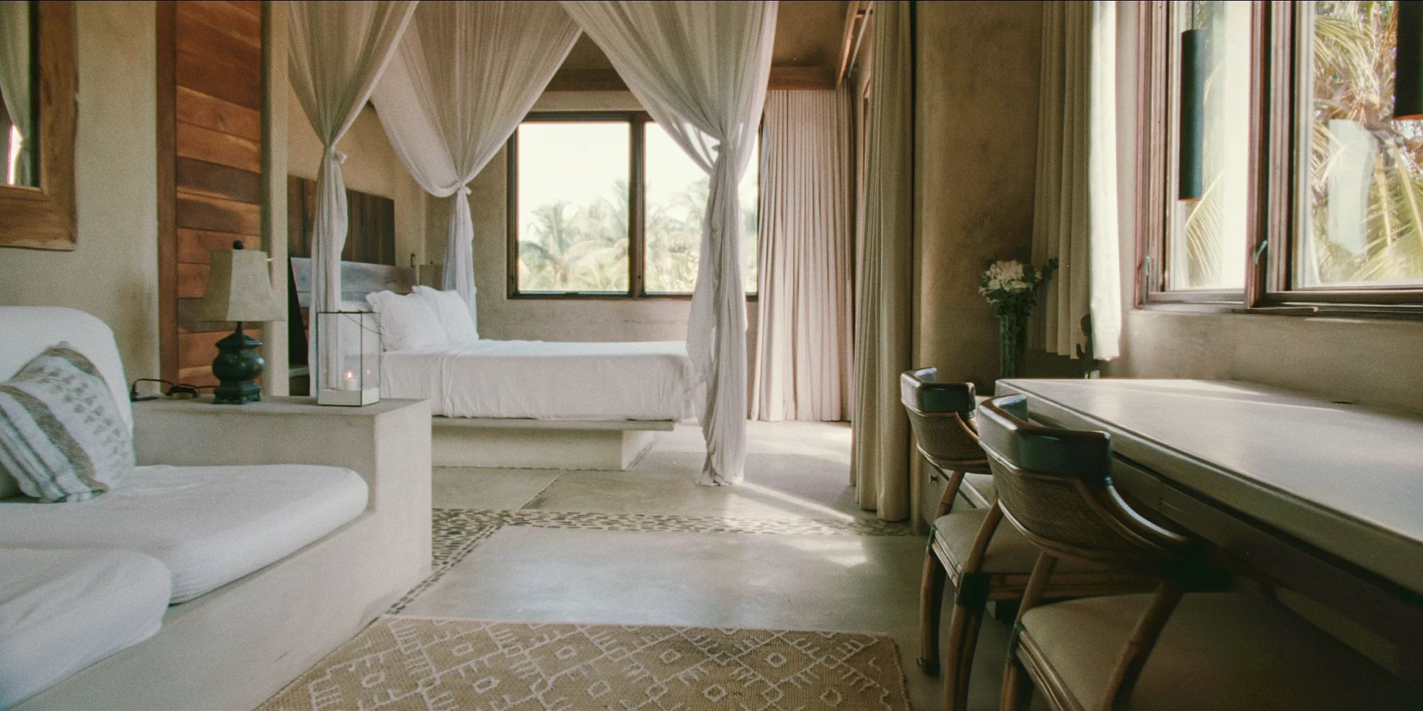 Interior of a cozy bedroom with a white canopy bed near a window, a beige sofa with a pillow, a table lamp, a desk, and chairs, with tropical palm trees visible outside.