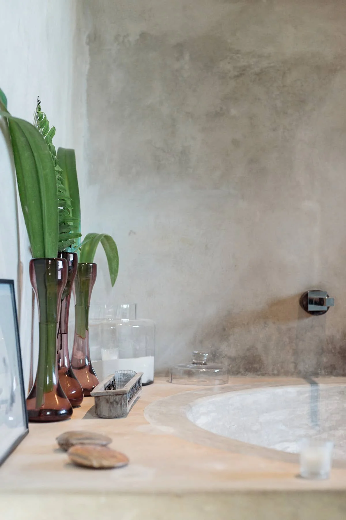 A bathroom countertop with three tall vases of green plants, two candles, a framed photograph, and small decorative rocks against a textured wall.