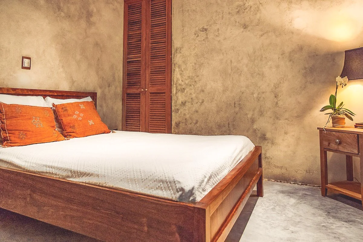 A bedroom with a wooden bed, white bedding, and orange pillows. There is a wooden side table with a lamp and a potted orchid. The walls are textured beige, and there is a wooden closet door.