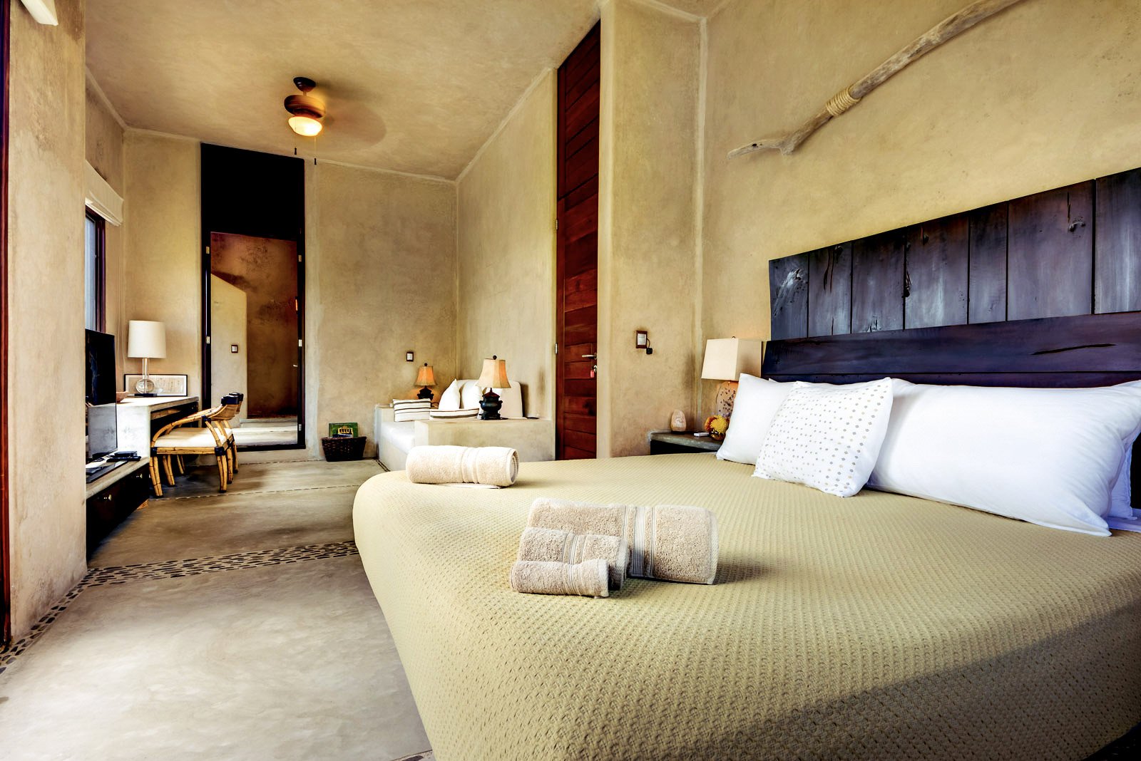 A cozy hotel room with a green bedspread, rolled towels, pillows, wooden headboard, bedside lamps, and a sitting area in the background.