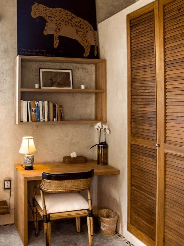 A cozy corner with a wooden desk, a wicker chair with a white cushion, a small table lamp, a tall vase with white orchids, and a wooden closet with louvered doors. There is a framed picture, books, and candles on a wall shelf above the desk, and a pi