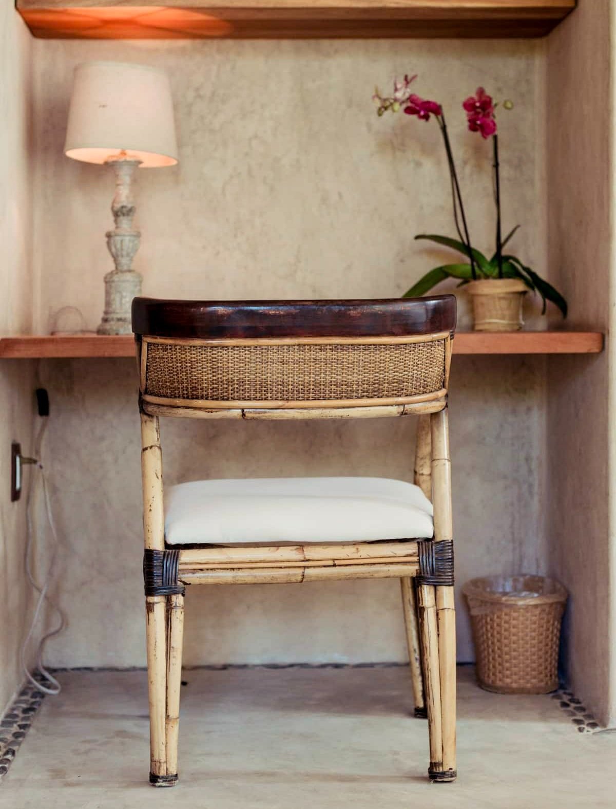 A cozy nook with a bamboo chair that has a cushioned seat, a wooden desk with a small lamp, a potted orchid plant, a wicker trash bin, and a wooden shelf against a beige wall.