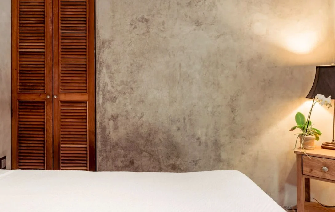 A bedroom corner with a wooden closet, a beige textured wall, a nightstand with a black lampshade, and a potted orchid plant.
