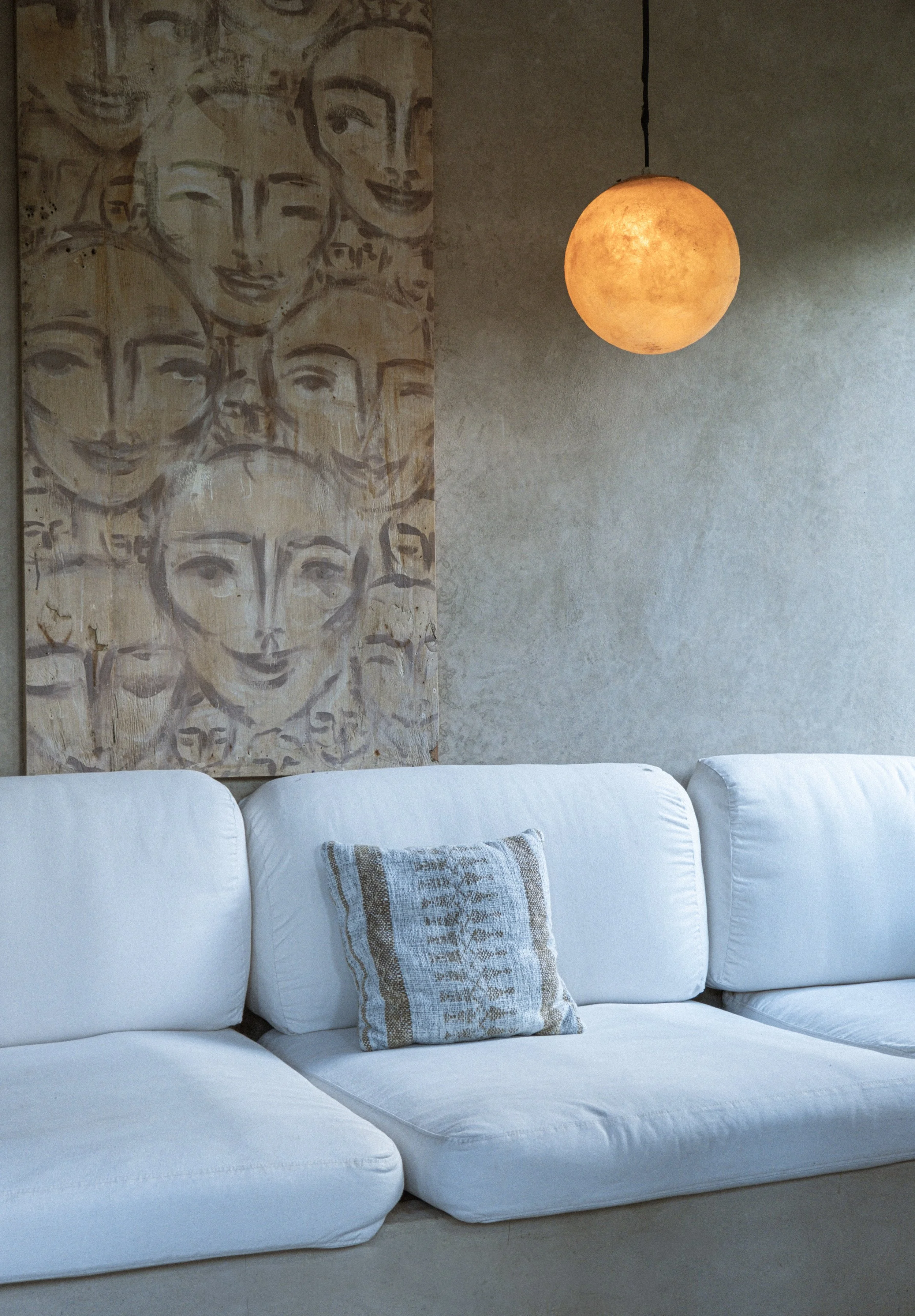 A white sofa with a patterned pillow sits in front of a textured wall decorated with a panel of smiling faces and a round orange hanging light.